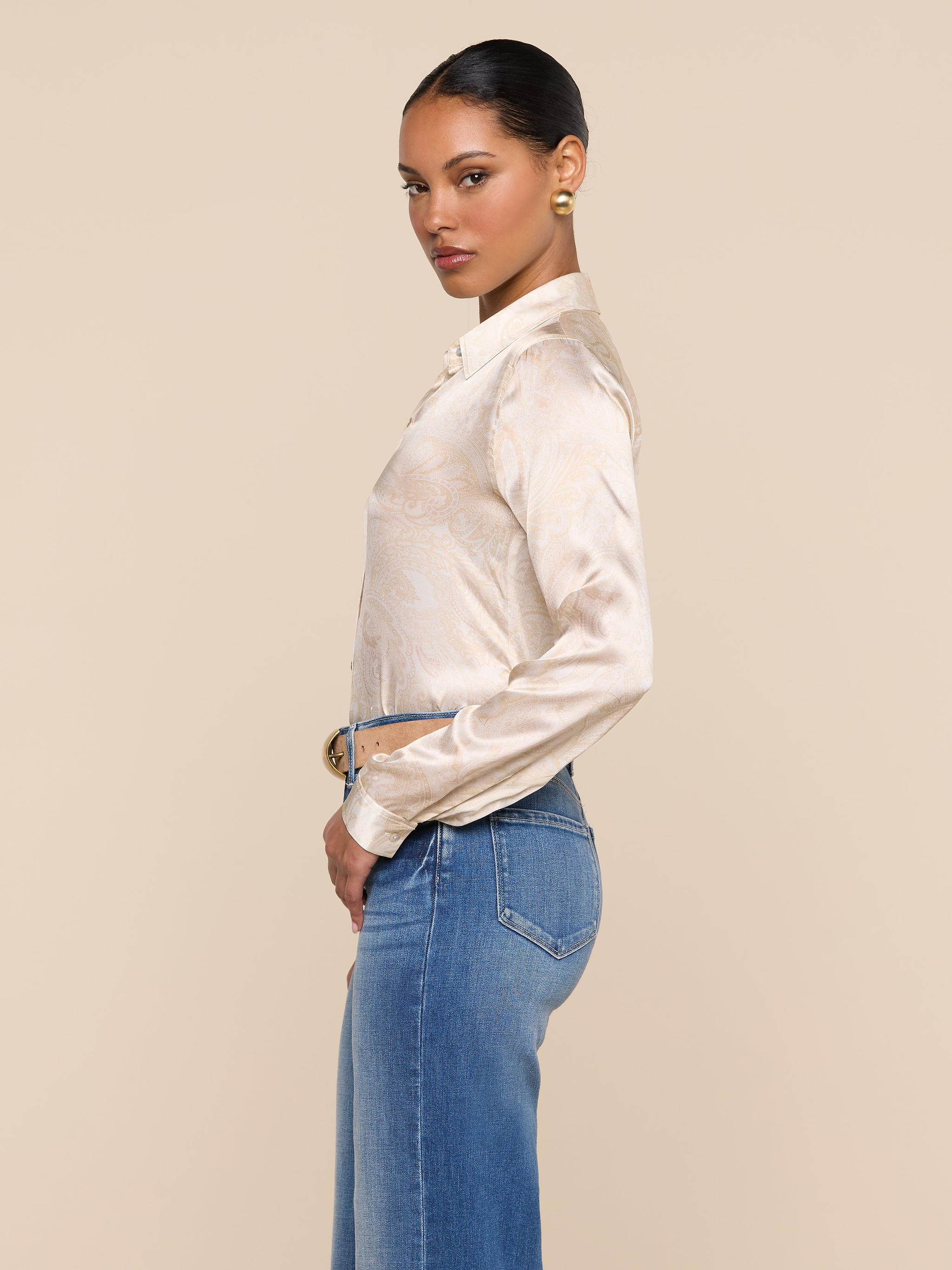 A woman stands in profile against a plain background, wearing the beige Tyler Silk Blouse with long sleeves, blue high-waisted jeans, and gold earrings. Her hair is styled in a neat bun.