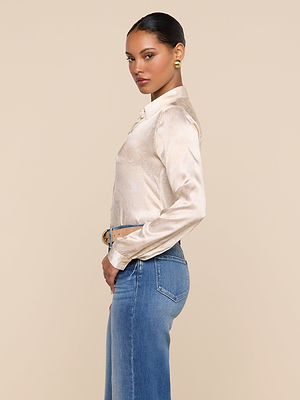 A woman with slicked-back hair and gold earrings wears the Tyler Silk Blouse—featuring a light paisley print—and blue jeans, standing in profile against a plain beige background while looking over her shoulder at the camera.
