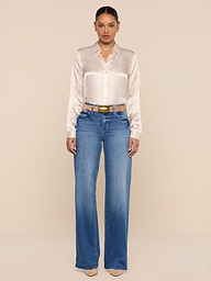 A woman stands facing forward, wearing the Tyler Silk Blouse from L’AGENCE tucked into high-waisted blue jeans and beige heels, against a plain white background.