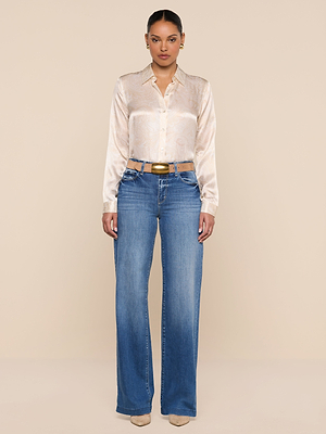 A woman stands facing forward, wearing the Tyler Silk Blouse with a paisley print, tucked into high-waisted, wide-leg blue jeans featuring hip cut-outs. She pairs the look with beige heels and a gold belt against a plain beige background.