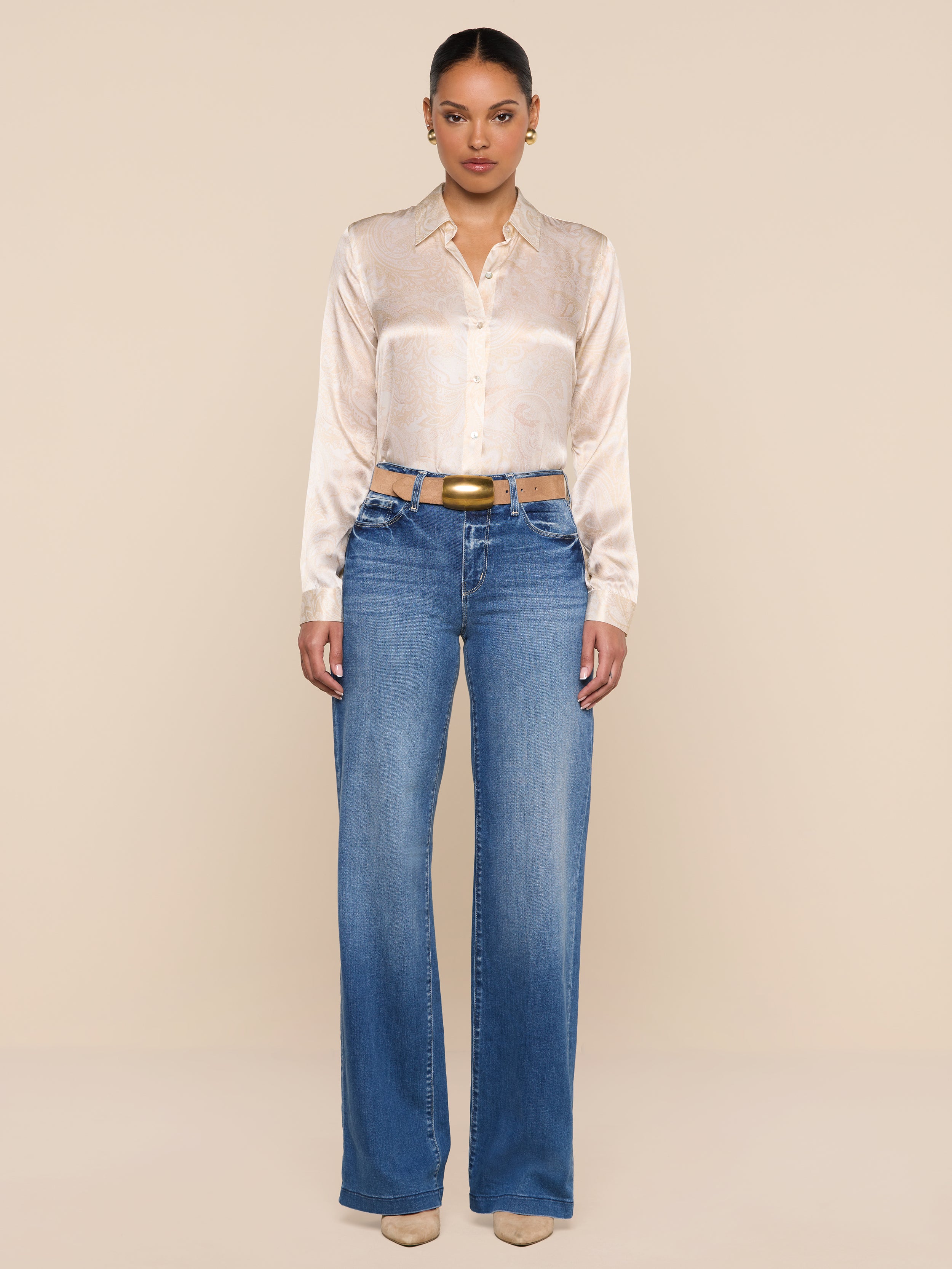 A woman stands facing forward, wearing the Tyler Silk Blouse from L’AGENCE tucked into high-waisted blue jeans and beige heels, against a plain white background.