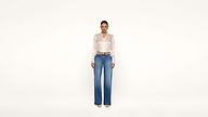 A woman stands against a plain white background wearing a shiny beige blouse, blue high-waisted wide-leg jeans, a brown belt, and beige pointed shoes.