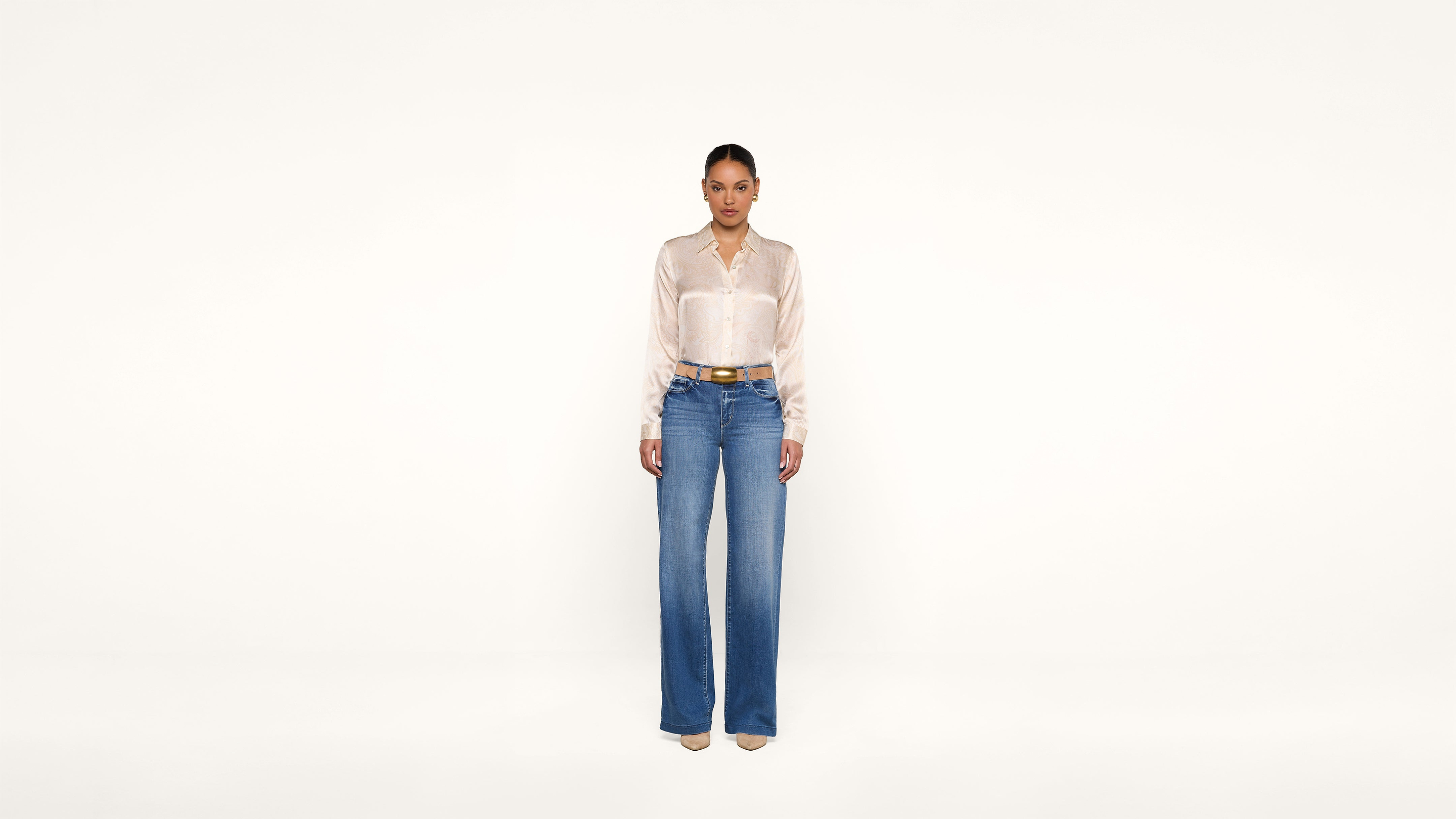 A woman stands against a plain white background wearing a shiny beige blouse, blue high-waisted wide-leg jeans, a brown belt, and beige pointed shoes.