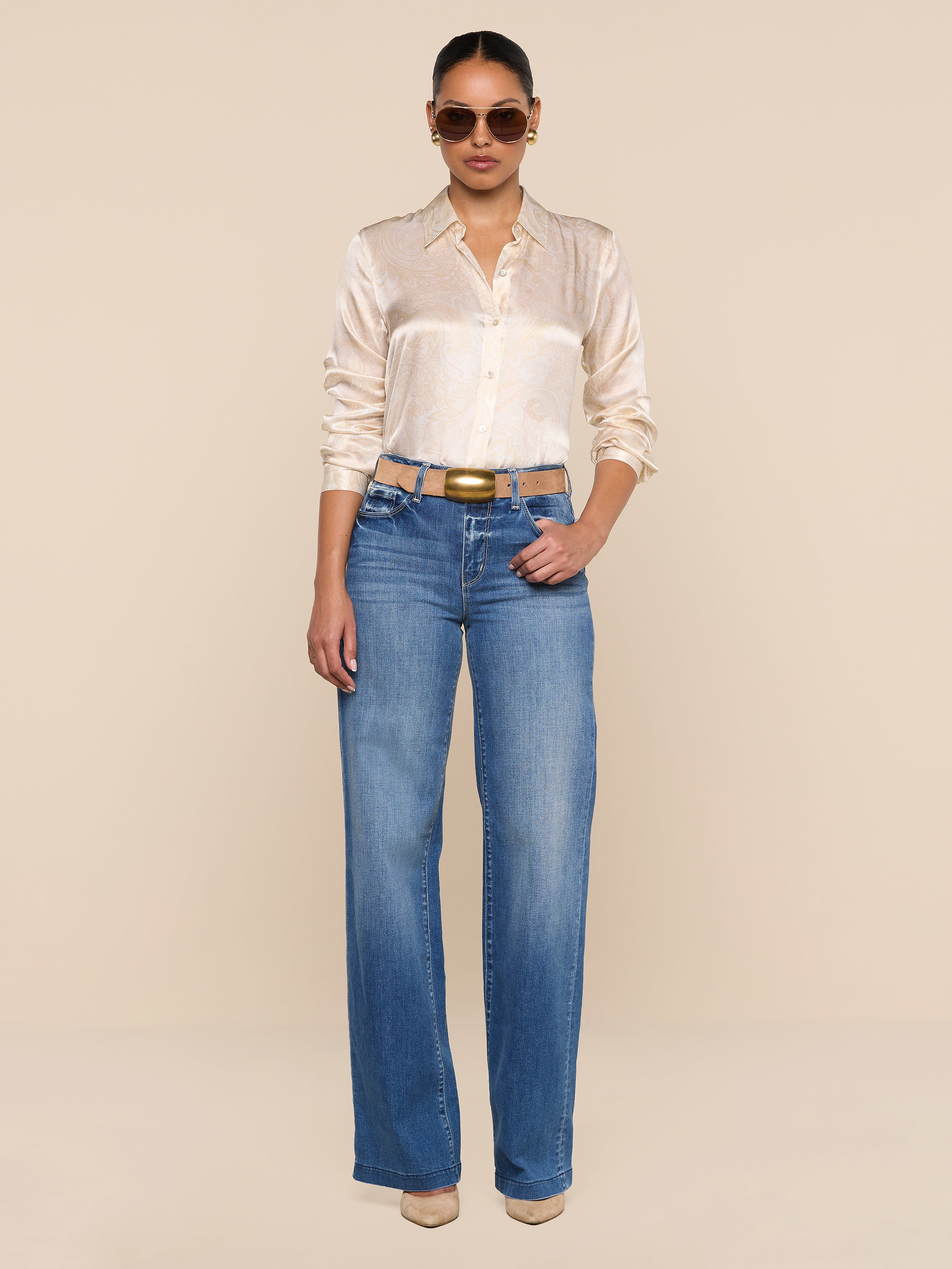A person wearing sunglasses, the Tyler Silk Blouse in a paisley print, blue wide-leg jeans, and beige heels stands against a plain white background.