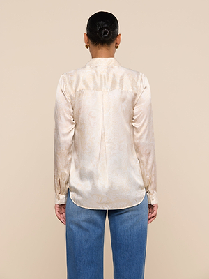 A person stands with their back to the camera, wearing the Tyler Silk Blouse—a light-colored, long-sleeve silk button-down with a subtle paisley print—and blue jeans, set against a plain beige background.