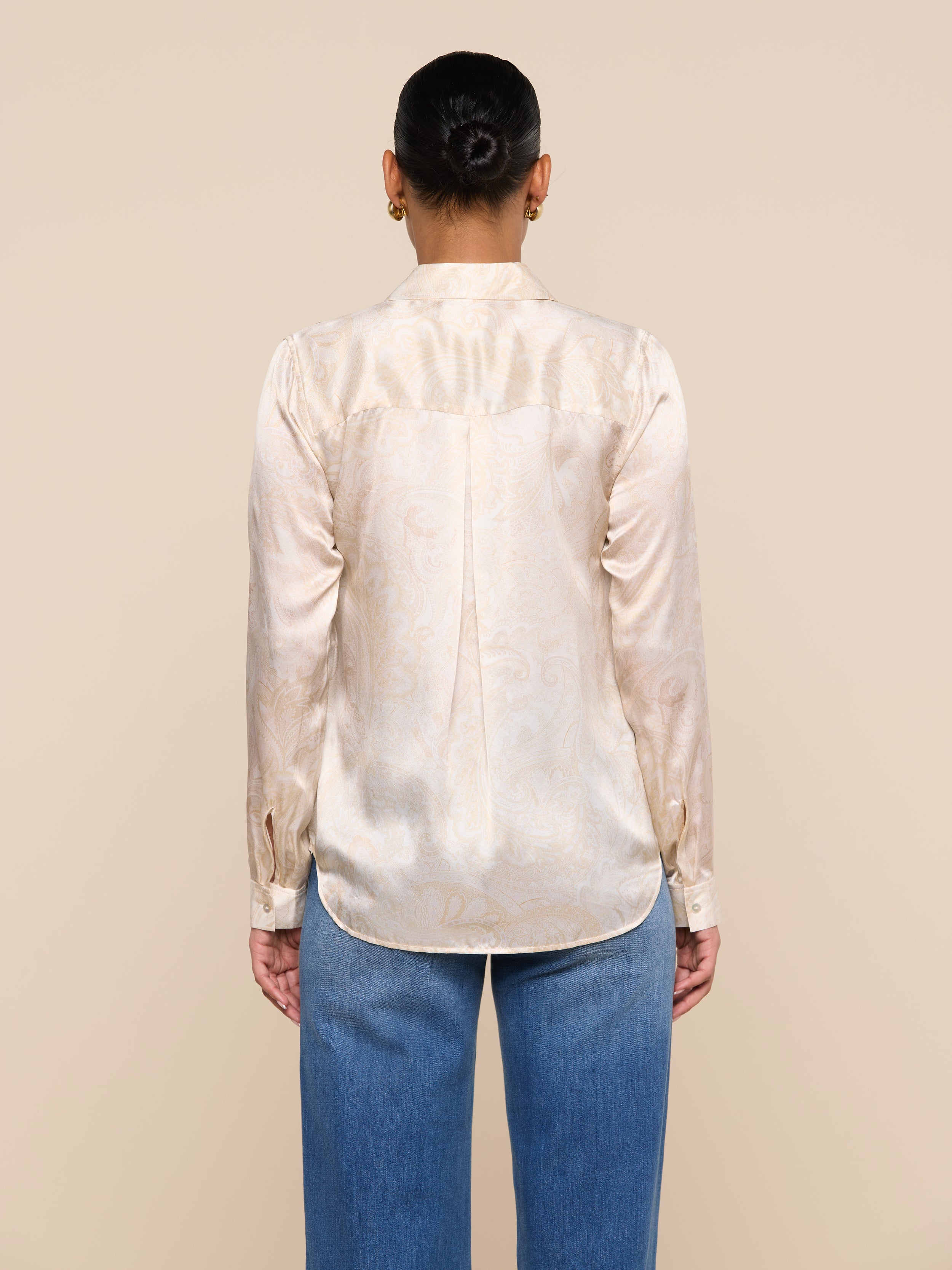 A person stands facing away from the camera, wearing the Tyler Silk Blouse—a cream-colored satin top with a subtle paisley print—paired with blue jeans, against a plain light background.