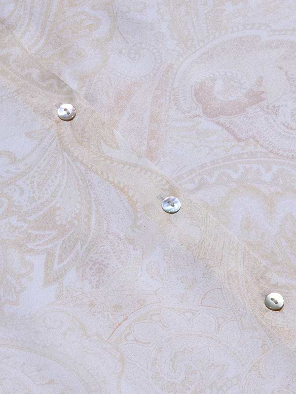 Close-up of the pale, paisley-patterned silk fabric of the Tyler Silk Blouse, featuring three iridescent buttons arranged diagonally. The soft beige and cream tones are adorned with subtle floral and leaf motifs, capturing L’AGENCE’s signature elegance.