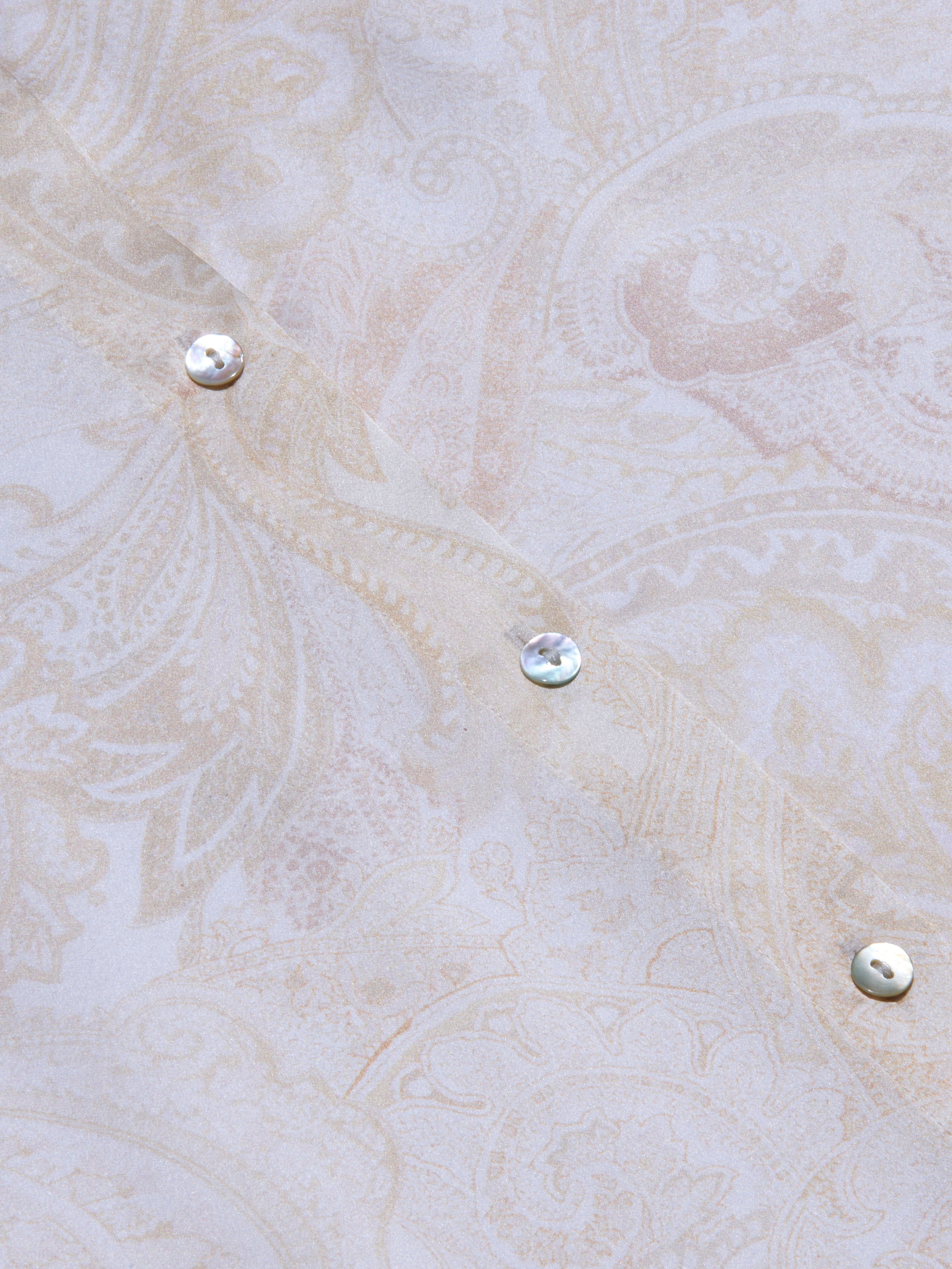 Close-up of the pale, paisley-patterned silk fabric of the Tyler Silk Blouse, featuring three iridescent buttons arranged diagonally. The soft beige and cream tones are adorned with subtle floral and leaf motifs, capturing L’AGENCE’s signature elegance.