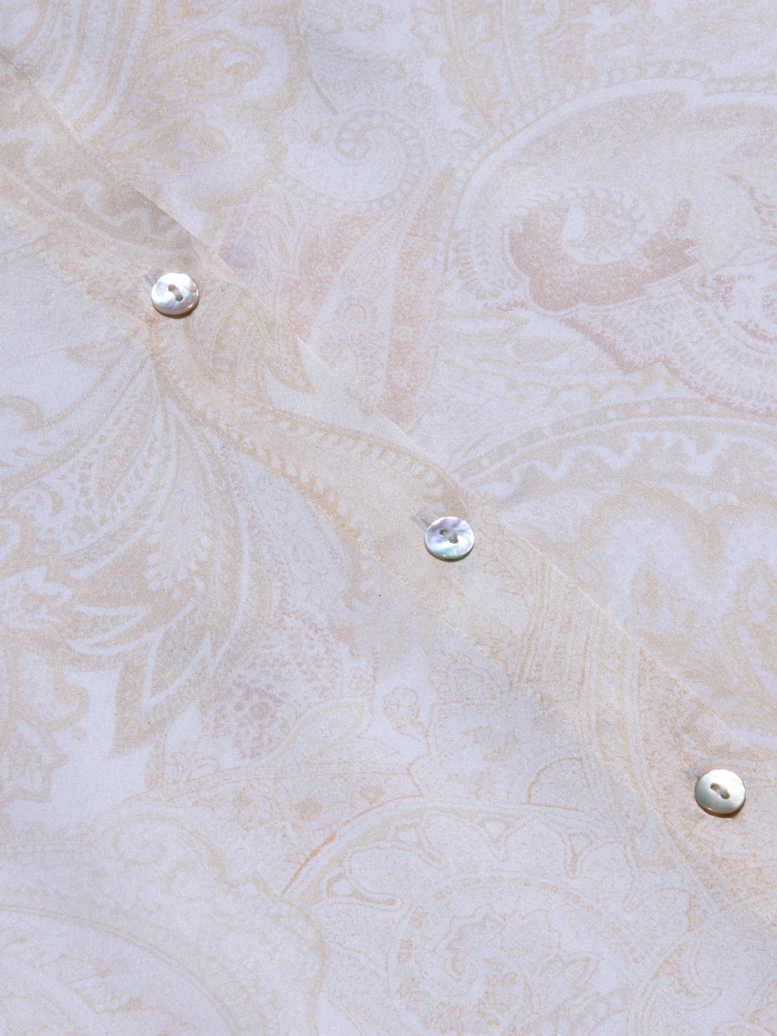 Close-up of the pale, paisley-patterned silk fabric of the Tyler Silk Blouse, featuring three iridescent buttons arranged diagonally. The soft beige and cream tones are adorned with subtle floral and leaf motifs, capturing L’AGENCE’s signature elegance.