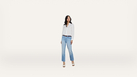 A woman stands on a light background, wearing a white button-up shirt, light blue jeans, a brown belt, and heeled sandals. She looks to her left with her hands by her sides.