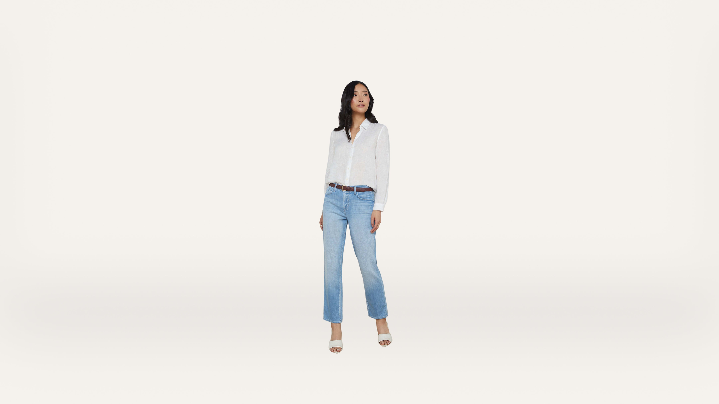 A woman stands on a light background, wearing a white button-up shirt, light blue jeans, a brown belt, and heeled sandals. She looks to her left with her hands by her sides.