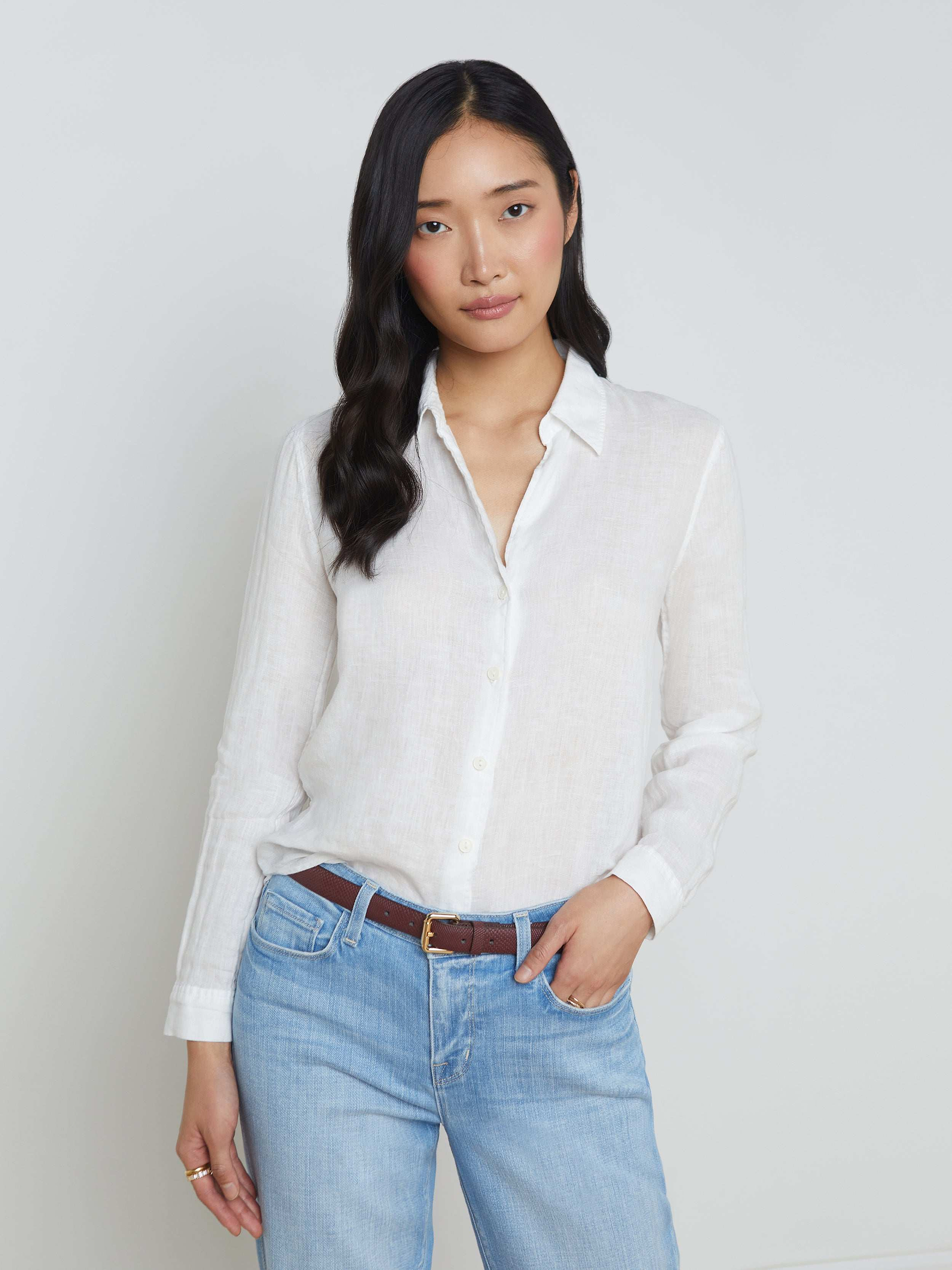 A person with long, wavy dark hair is wearing the Tyler Linen Blouse, a white long sleeve button-down shirt, paired with light blue jeans. They stand against a plain light background with one hand in their pocket and a relaxed expression.