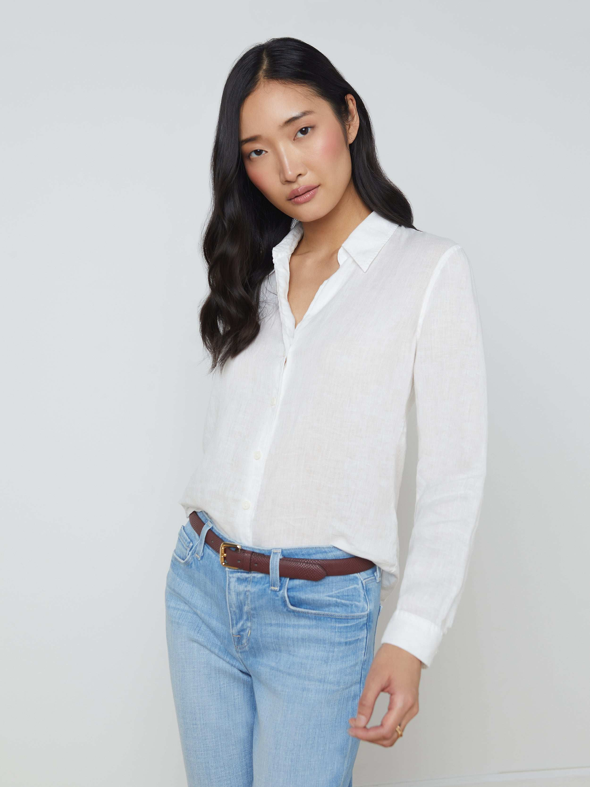 Woman with long black hair wearing the Tyler Linen Blouse, a white summer shirt, tucked into light blue jeans with a brown belt, standing against a plain light background.