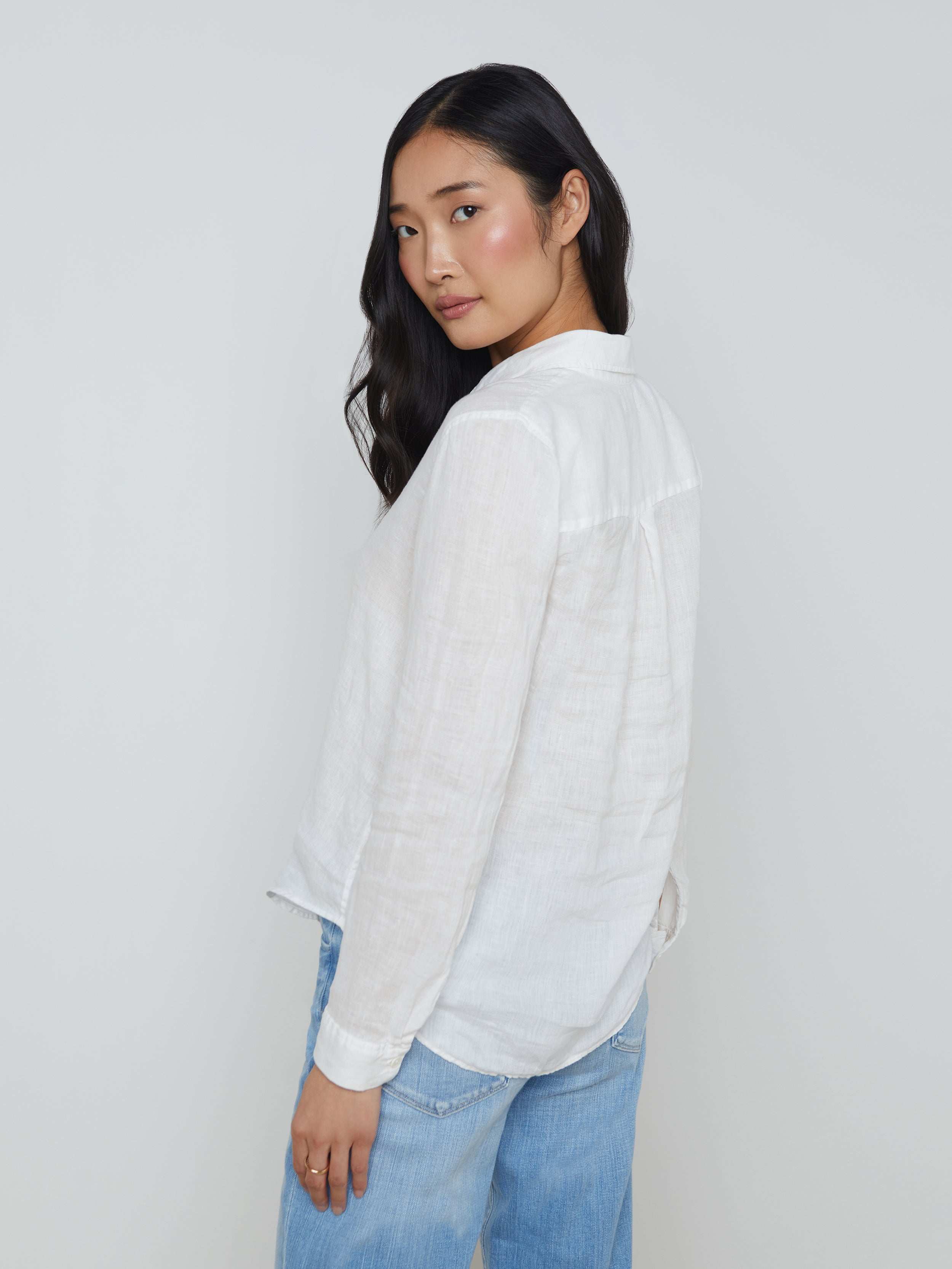 A woman with long dark hair, wearing the Tyler Linen Blouse—a white, long sleeve button-down shirt—and light blue jeans stands turned slightly to the side against a plain light background.