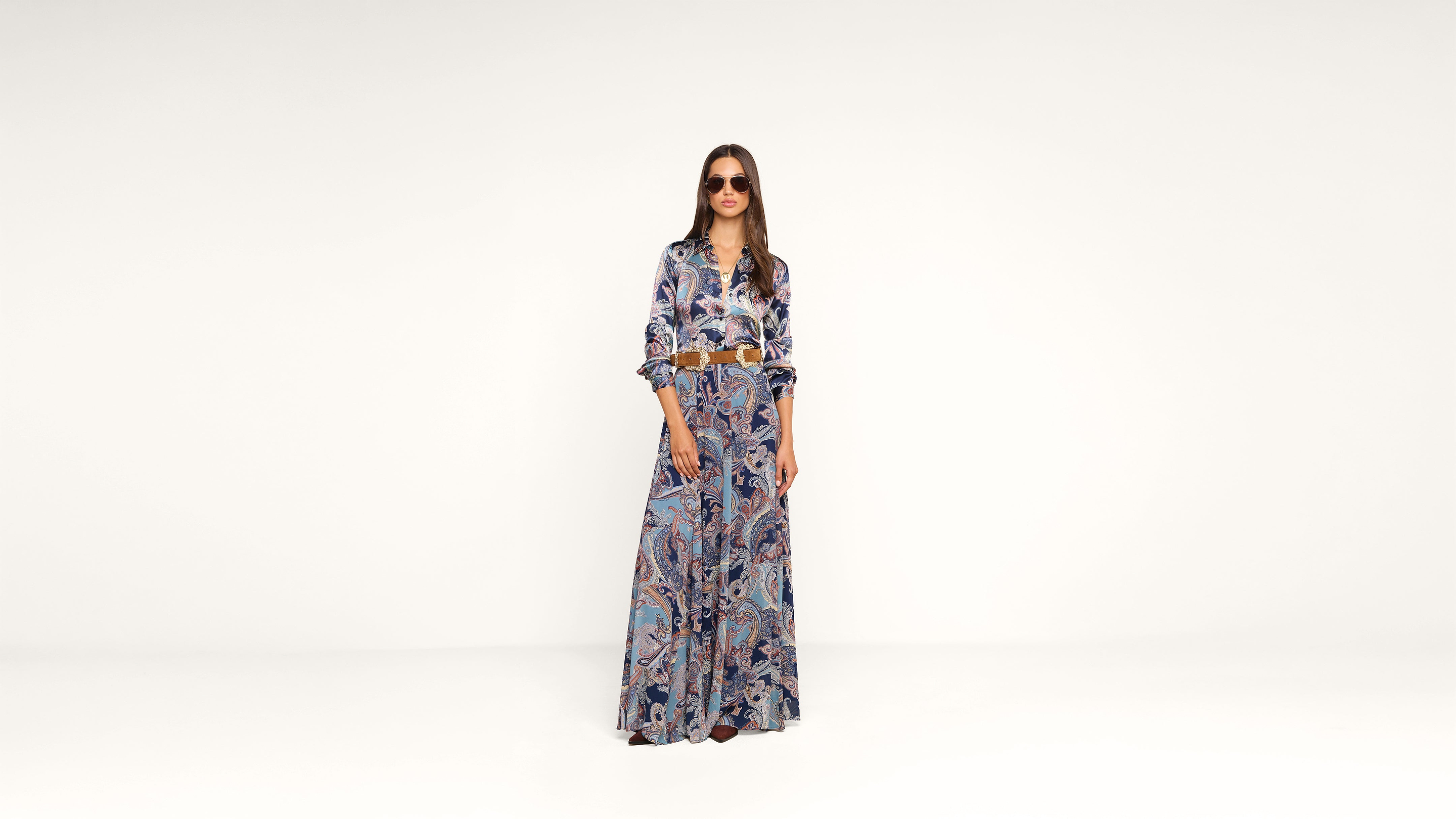 Woman wearing sunglasses and a patterned, button-up, long-sleeve shirt with a matching maxi skirt, standing against a plain white background.