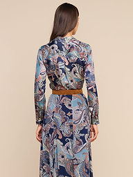 Une femme, de dos, porte le chemisier « Tyler Silk » à imprimé paisley bleu, agrémenté dune ceinture marron. Ses cheveux bruns et raides sont lâchés et se détachent sur un fond beige.