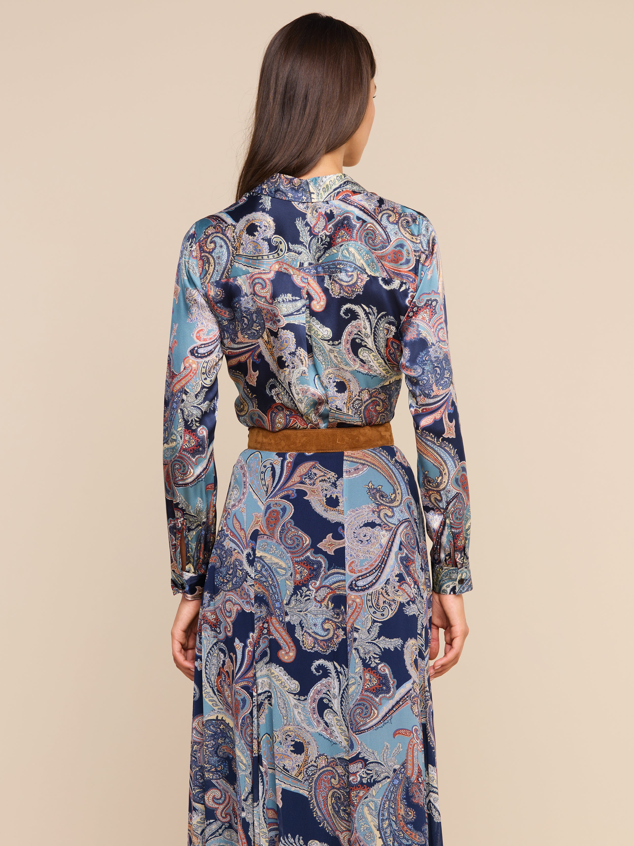 Une femme, de dos, porte le chemisier « Tyler Silk » à imprimé paisley bleu, agrémenté dune ceinture marron. Ses cheveux bruns et raides sont lâchés et se détachent sur un fond beige.
