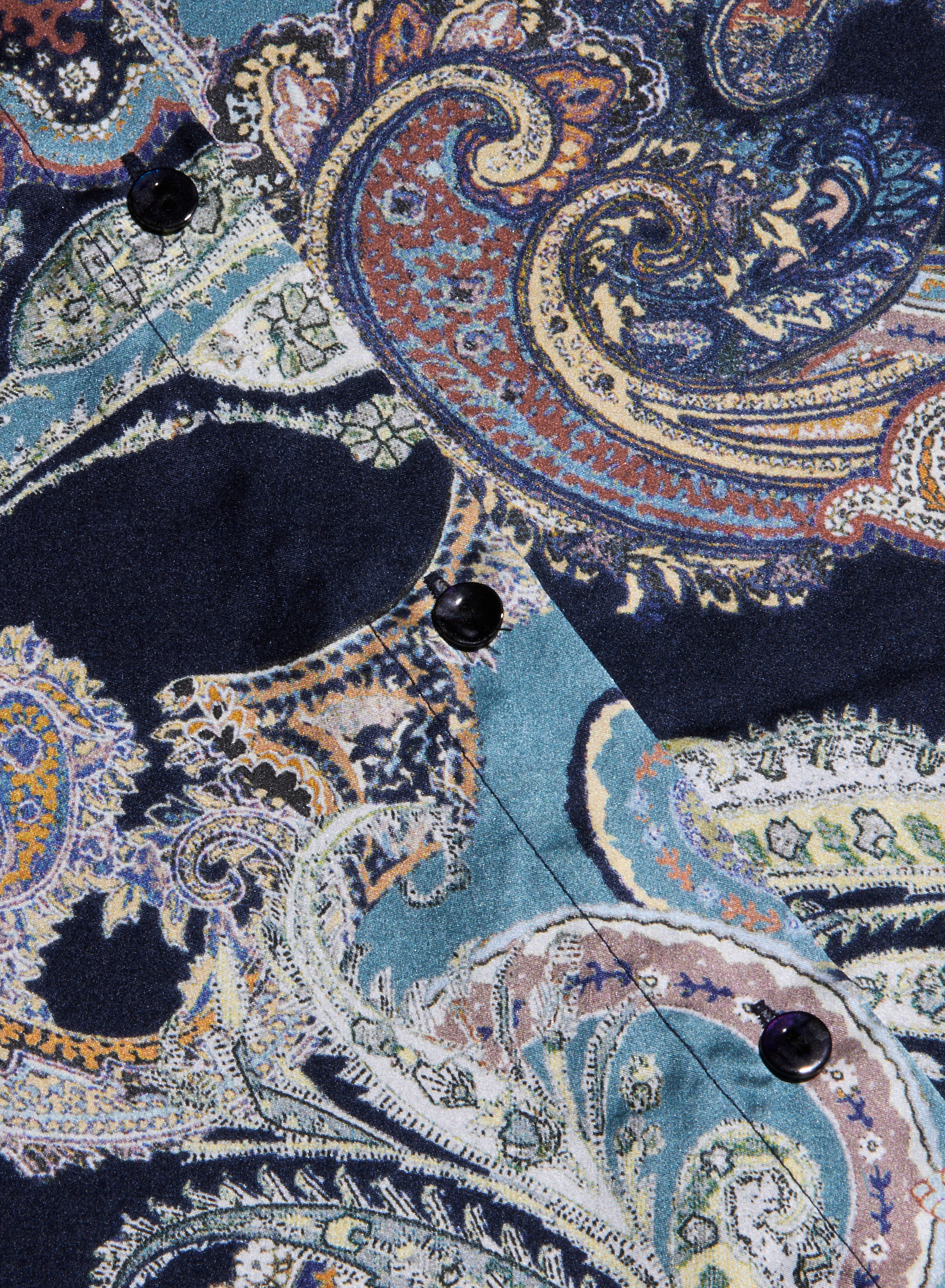 Close-up of the Tyler Silk Blouse fabric featuring a colorful, intricate paisley print in shades of blue, navy, gold, and teal, with black buttons accenting a stitched seam.