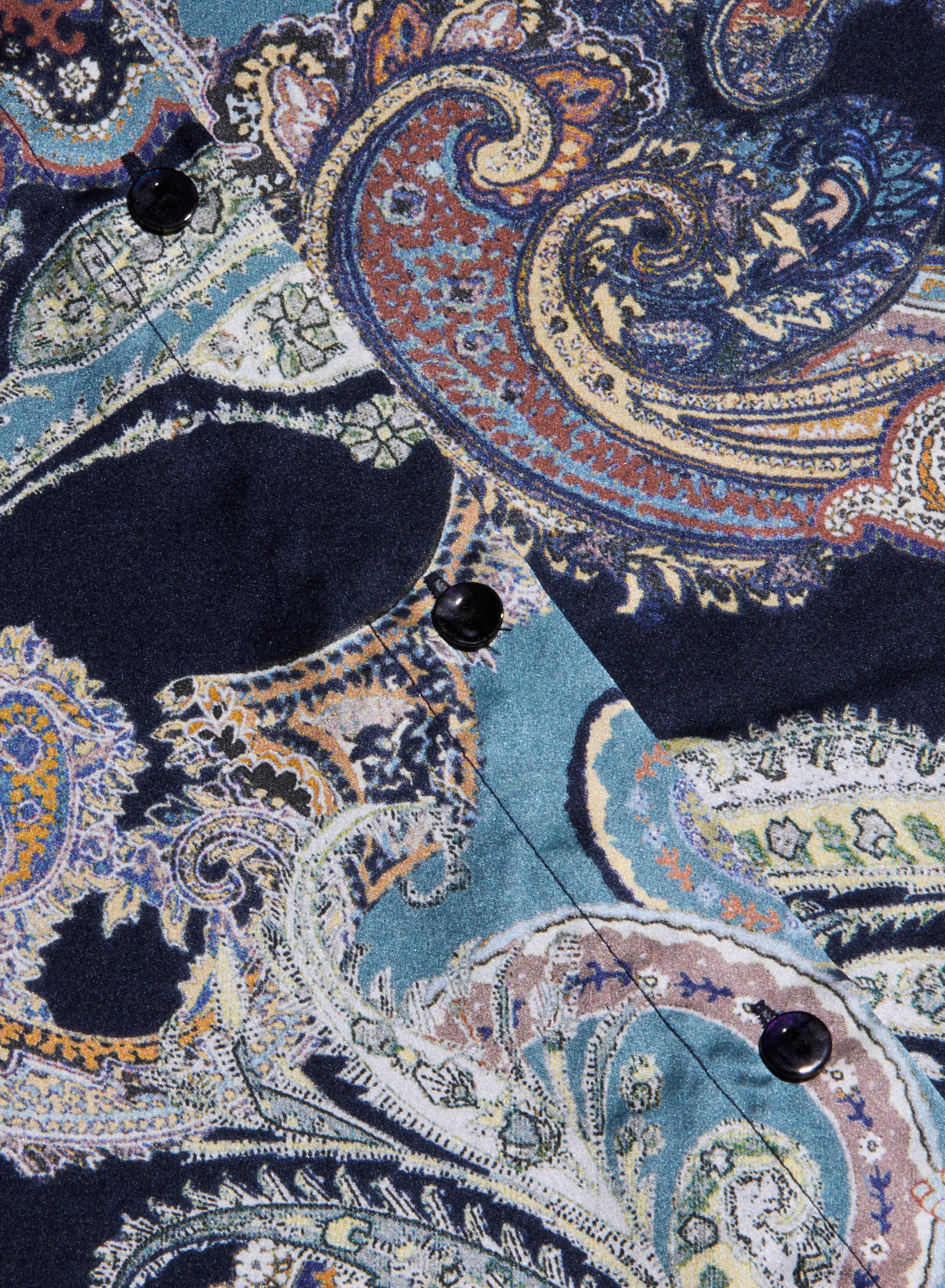 Close-up of the Tyler Silk Blouse fabric featuring a colorful, intricate paisley print in shades of blue, navy, gold, and teal, with black buttons accenting a stitched seam.