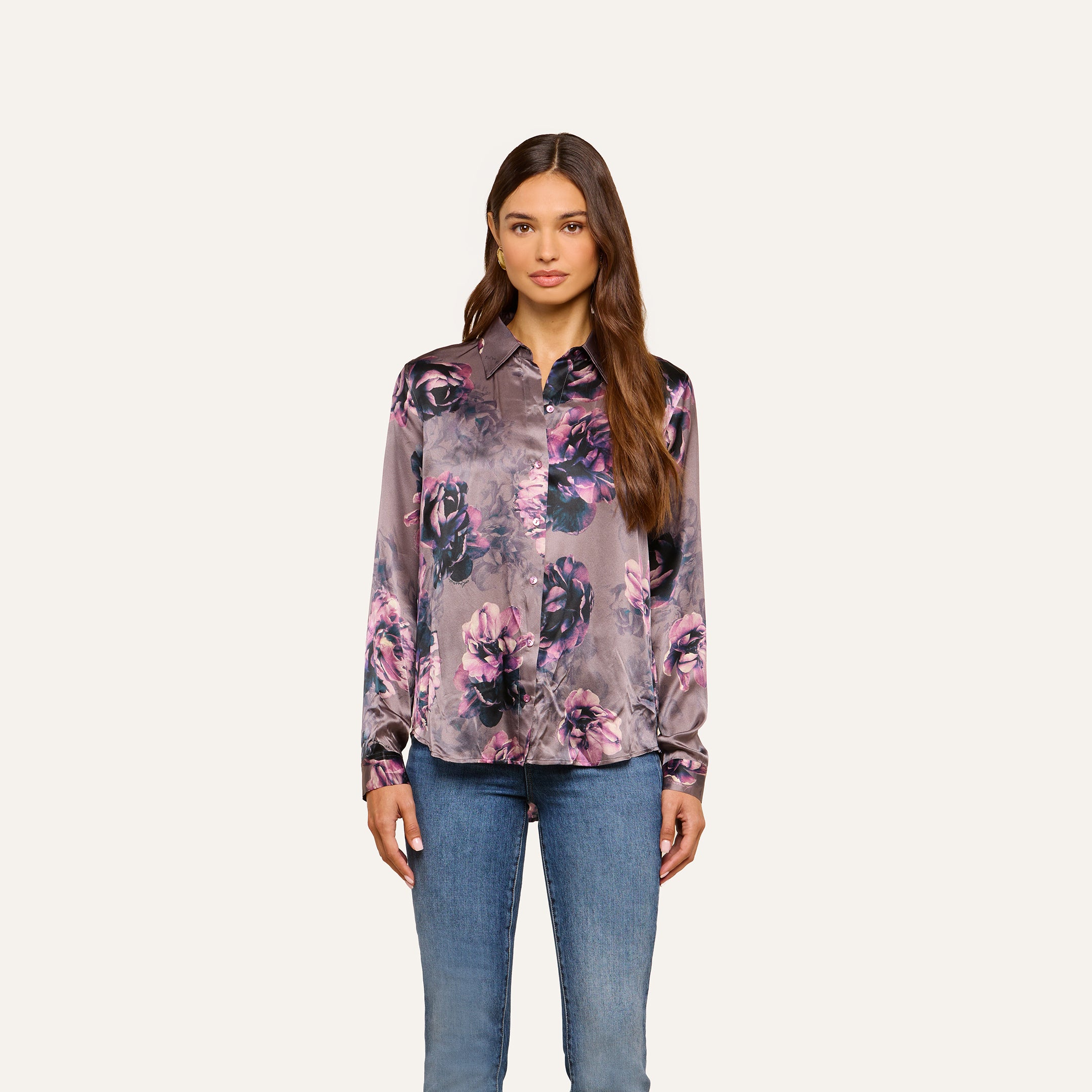 Woman with long brown hair wears the Tyler Silk Blouse by L’AGENCE in a purple floral print and blue jeans, standing against a plain, light background.