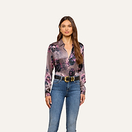 A woman stands facing forward, wearing a L’AGENCE Tyler Silk Blouse in purple floral print tucked into blue jeans with a black and gold belt, against a plain light background.