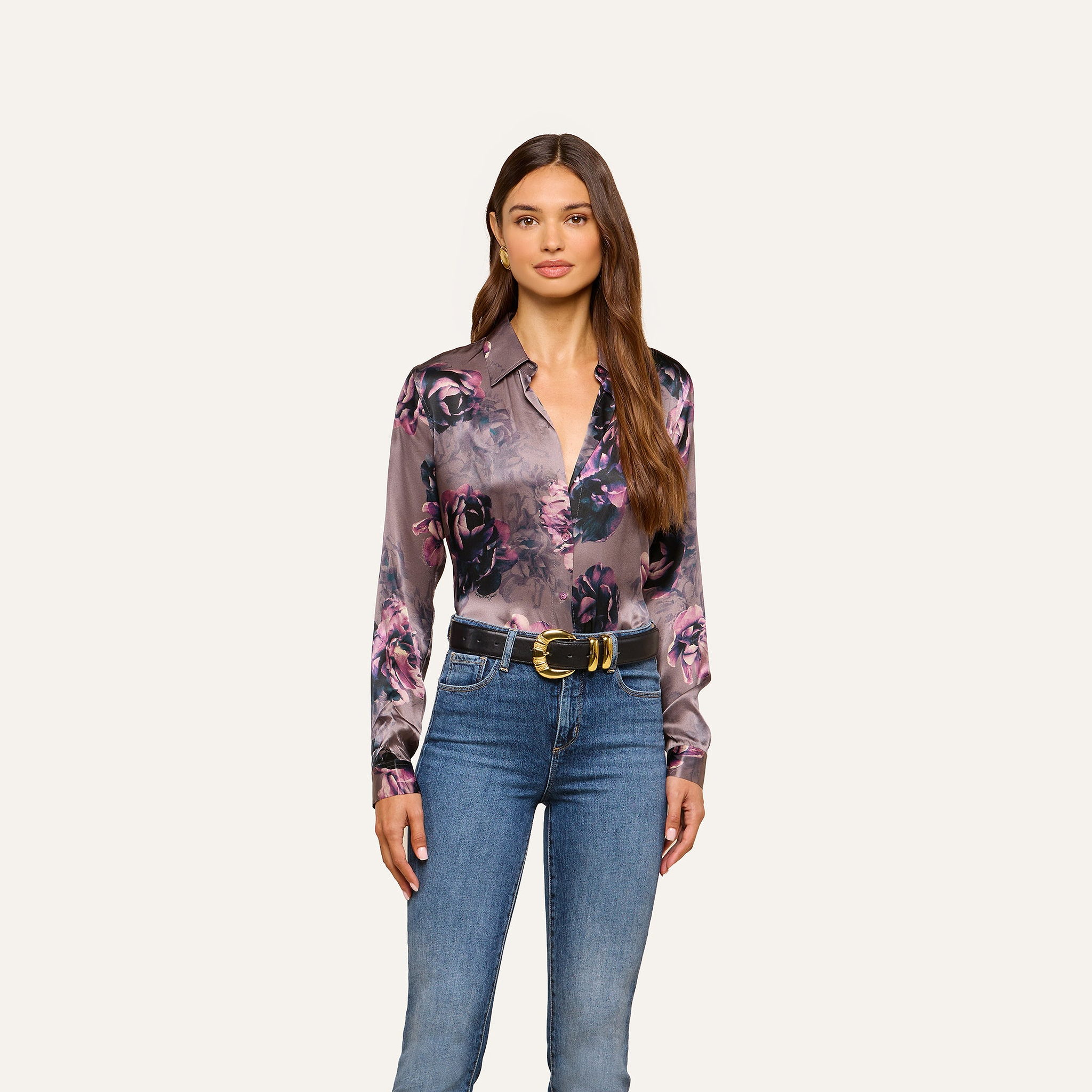 A woman stands facing forward, wearing a L’AGENCE Tyler Silk Blouse in purple floral print tucked into blue jeans with a black and gold belt, against a plain light background.
