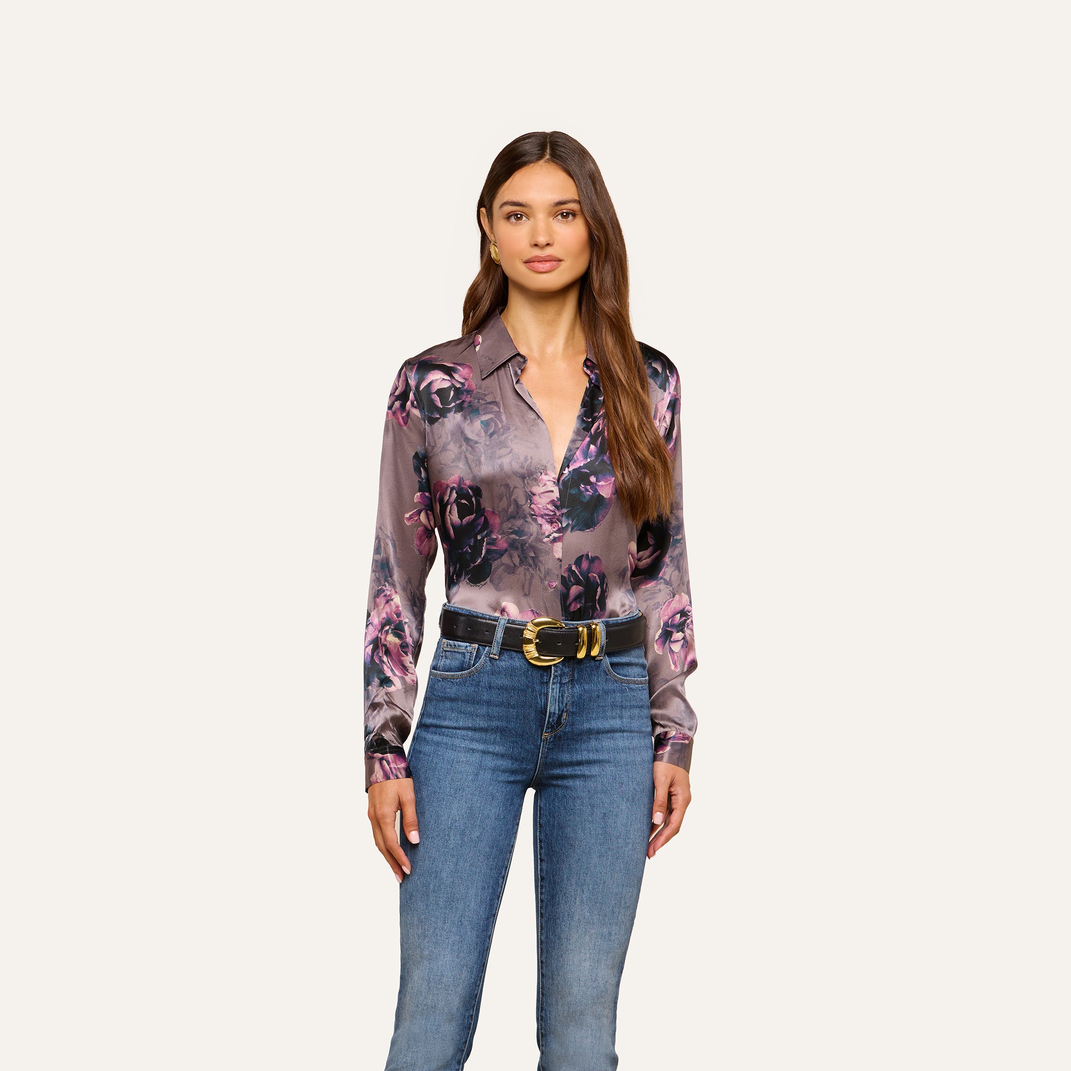 A woman stands facing forward, wearing a L’AGENCE Tyler Silk Blouse in purple floral print tucked into blue jeans with a black and gold belt, against a plain light background.