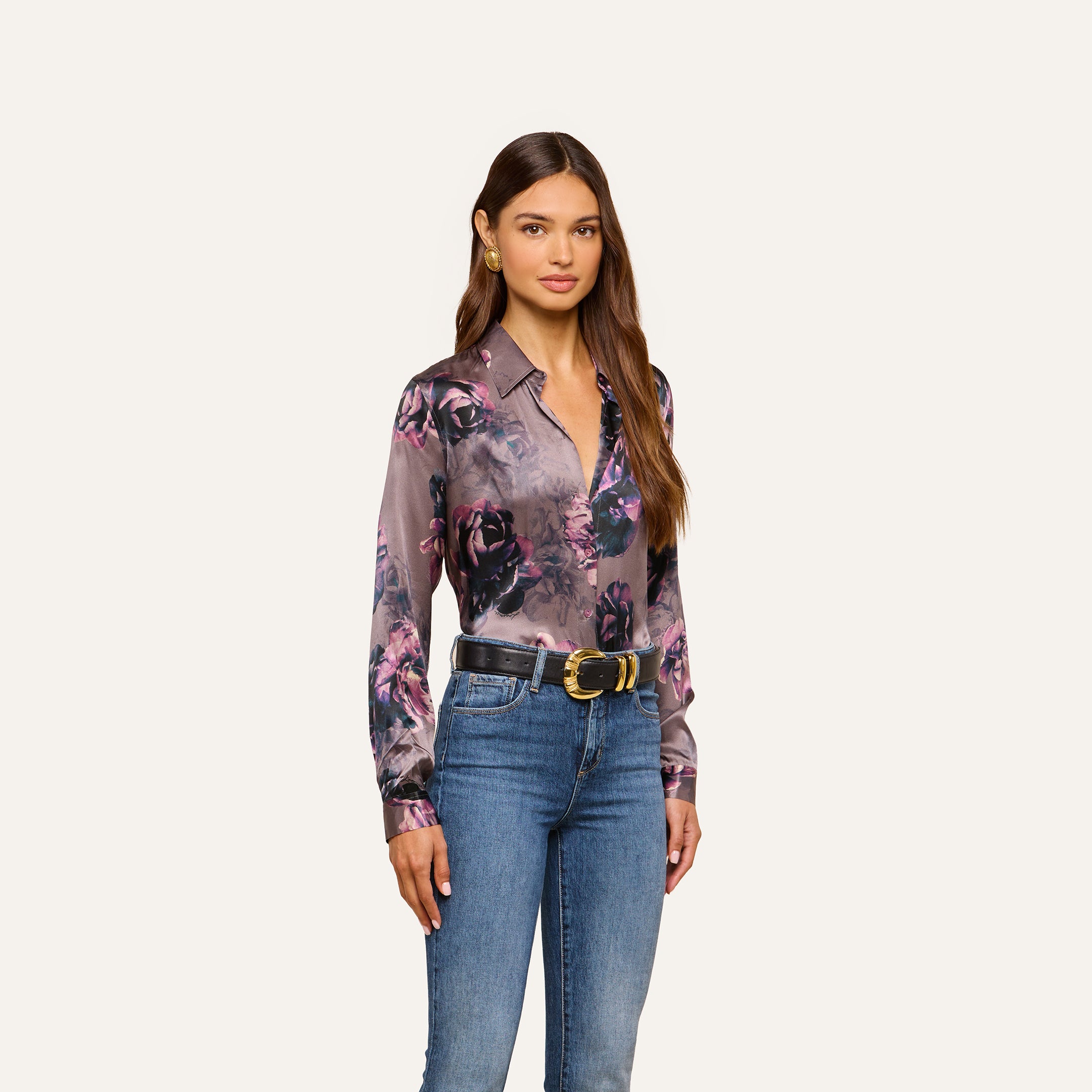 A woman stands against a light background, wearing the Tyler Silk Blouse—a purple floral print silk shirt from L’AGENCE—tucked into blue jeans with a black belt. She has long brown hair and a neutral expression.
