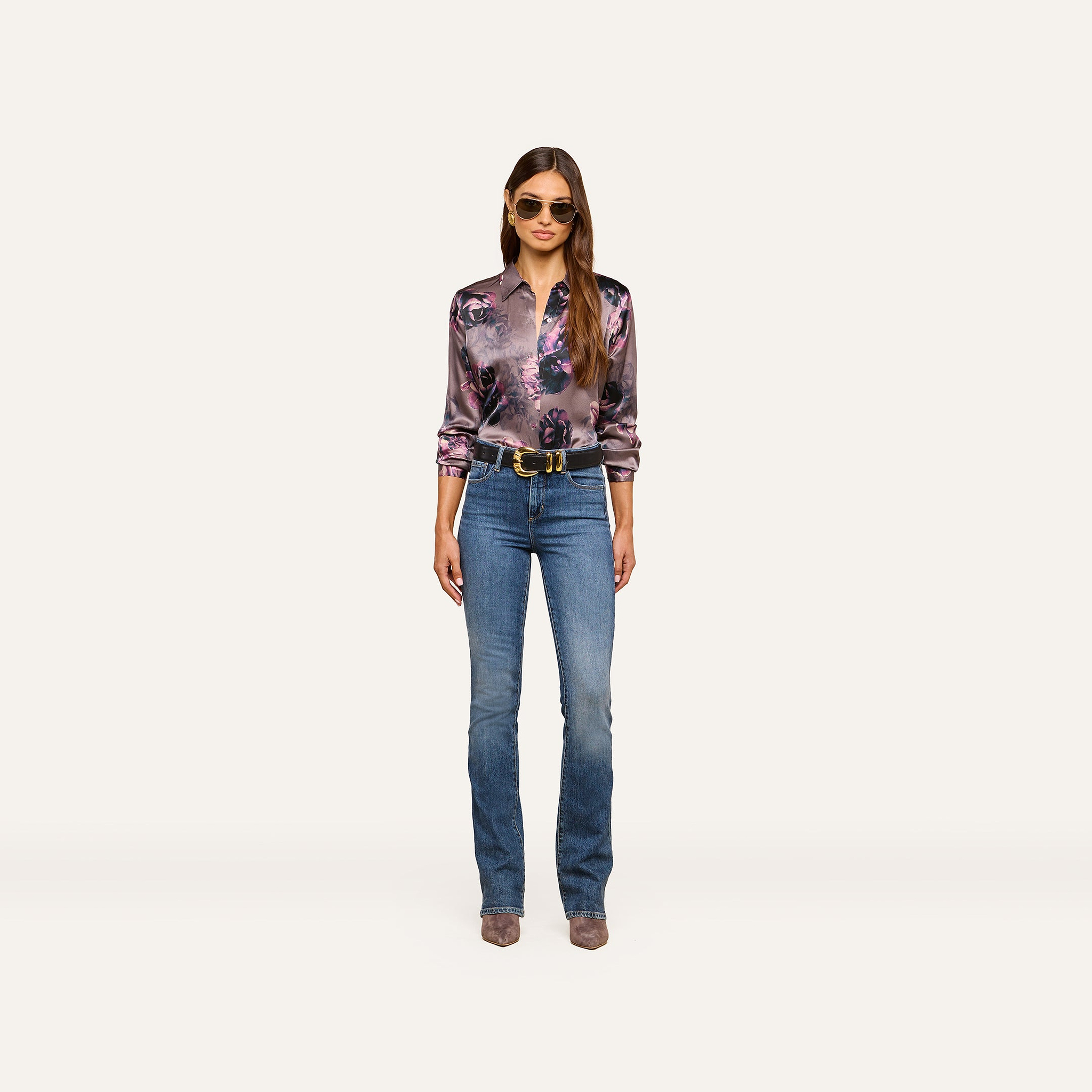A woman stands facing forward, wearing sunglasses, the Tyler Silk Blouse by L’AGENCE in a shiny purple floral print, blue jeans, a black belt, and heeled boots, with her long hair down against a plain light background.