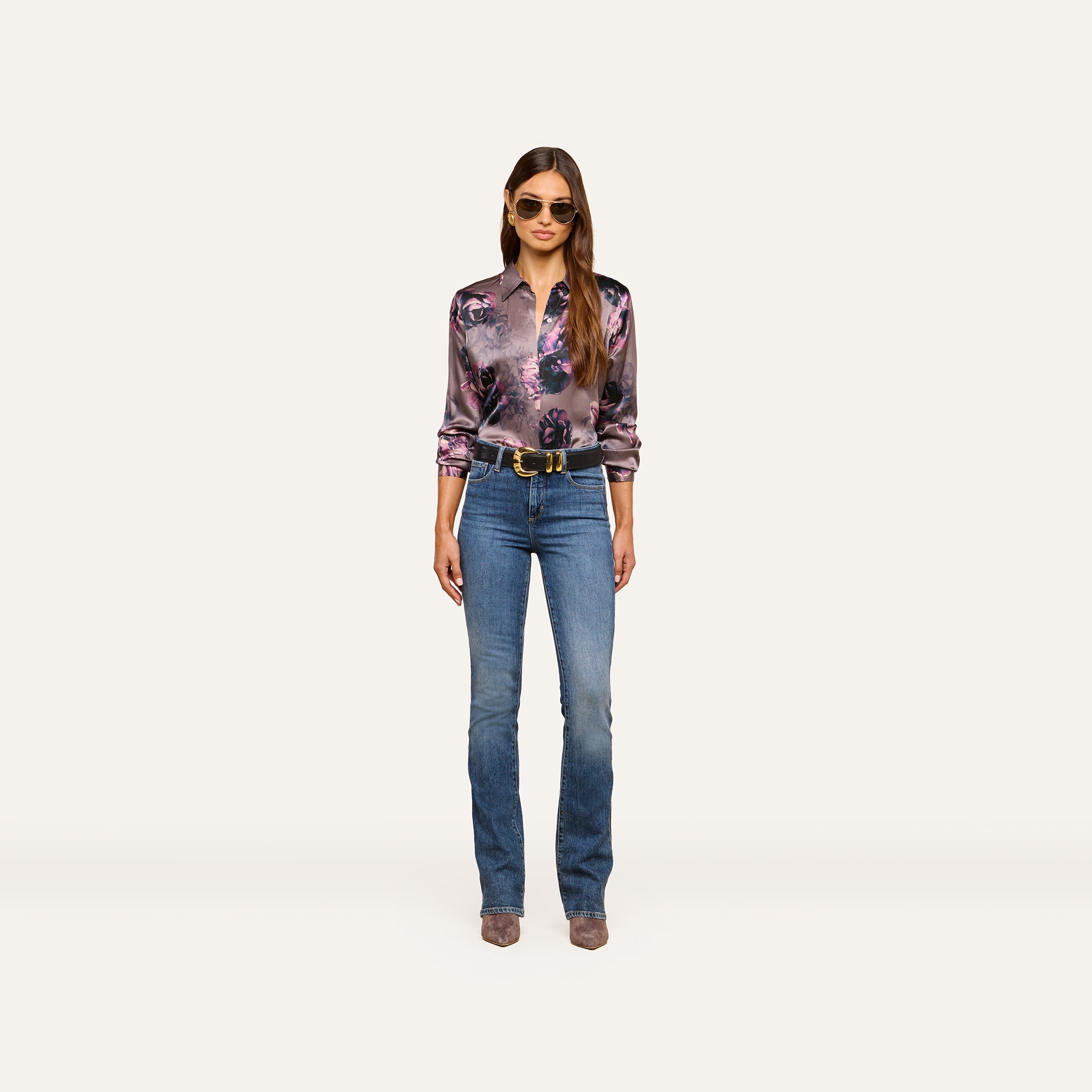 A woman stands facing forward, wearing sunglasses, the Tyler Silk Blouse by L’AGENCE in a shiny purple floral print, blue jeans, a black belt, and heeled boots, with her long hair down against a plain light background.