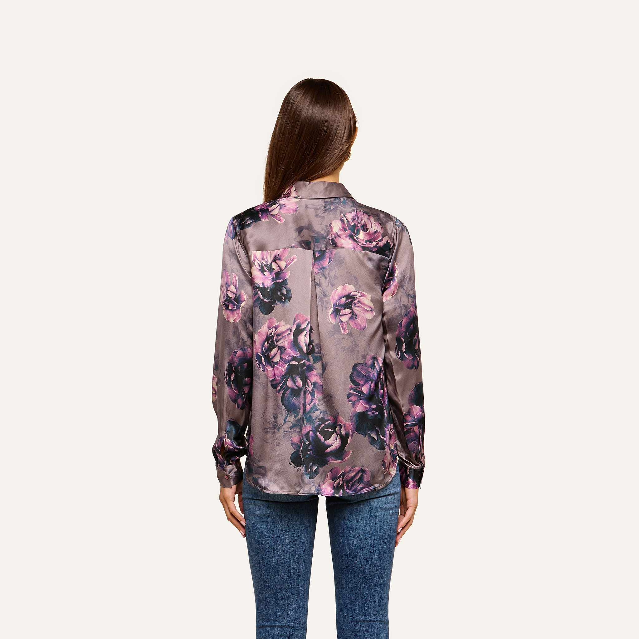 A woman with long brown hair, seen from the back, wears the Tyler Silk Blouse in a purple floral print paired with blue jeans, standing against a plain light background.