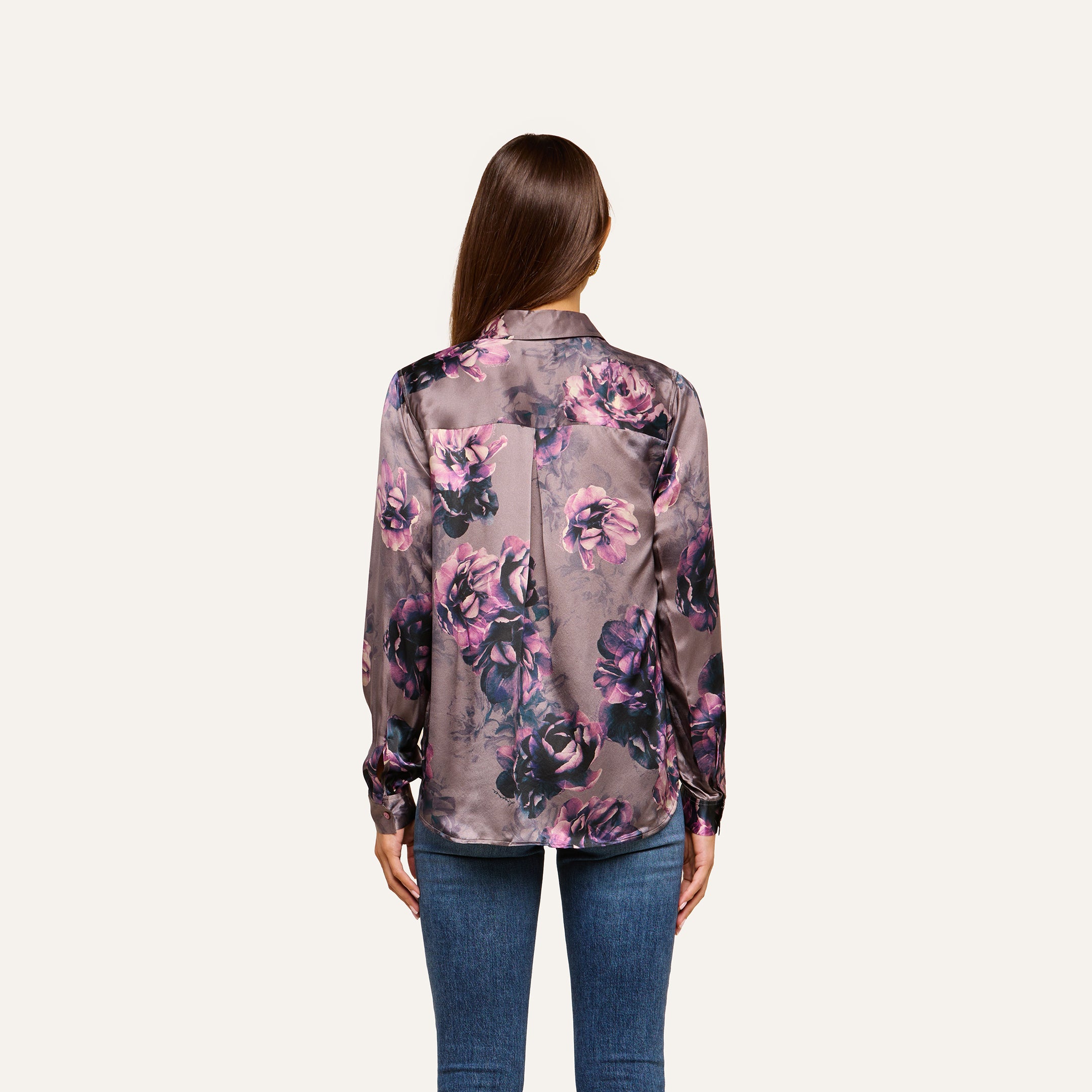 A woman with long brown hair, seen from the back, wears the Tyler Silk Blouse in a purple floral print paired with blue jeans, standing against a plain light background.