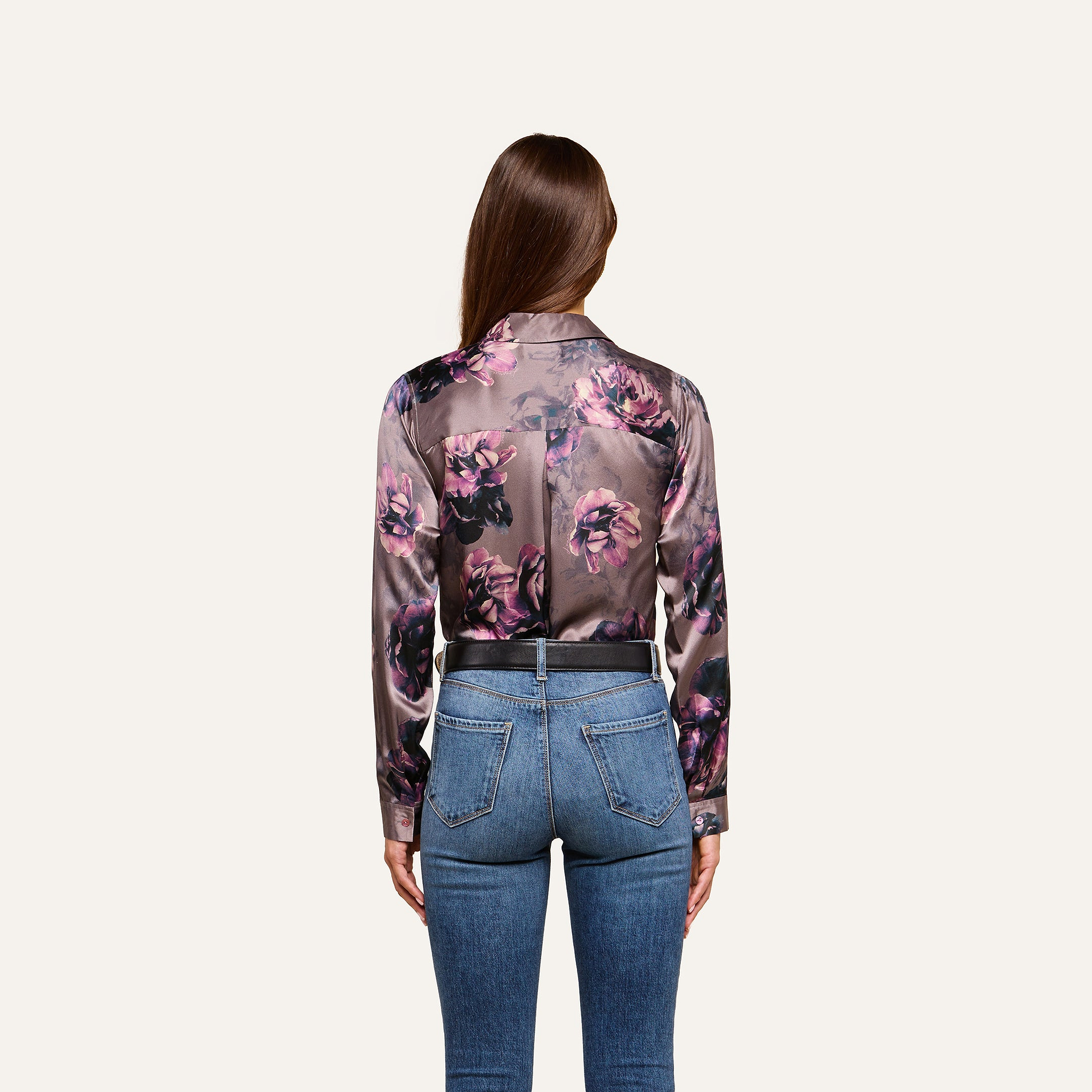 A woman with long brown hair, wearing the Tyler Silk Blouse in a purple floral print by L’AGENCE and blue high-waisted jeans, stands facing away from the camera against a plain light background.