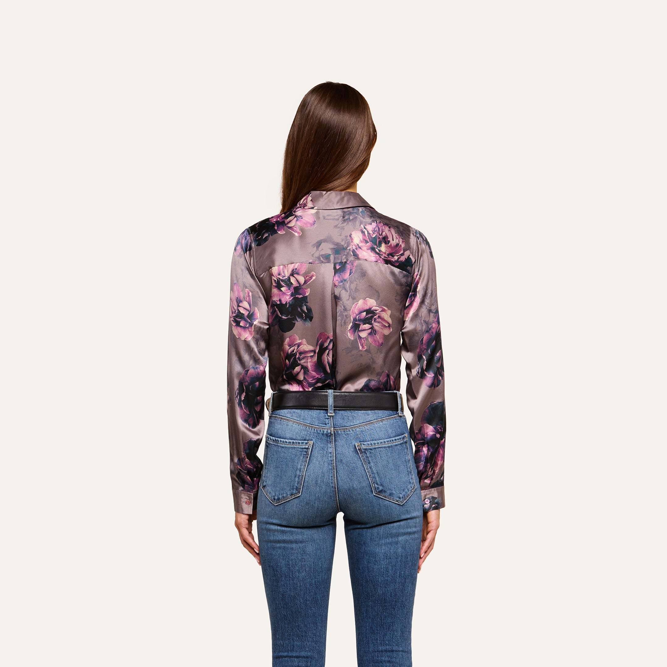 A woman with long brown hair, wearing the Tyler Silk Blouse in a purple floral print by L’AGENCE and blue high-waisted jeans, stands facing away from the camera against a plain light background.