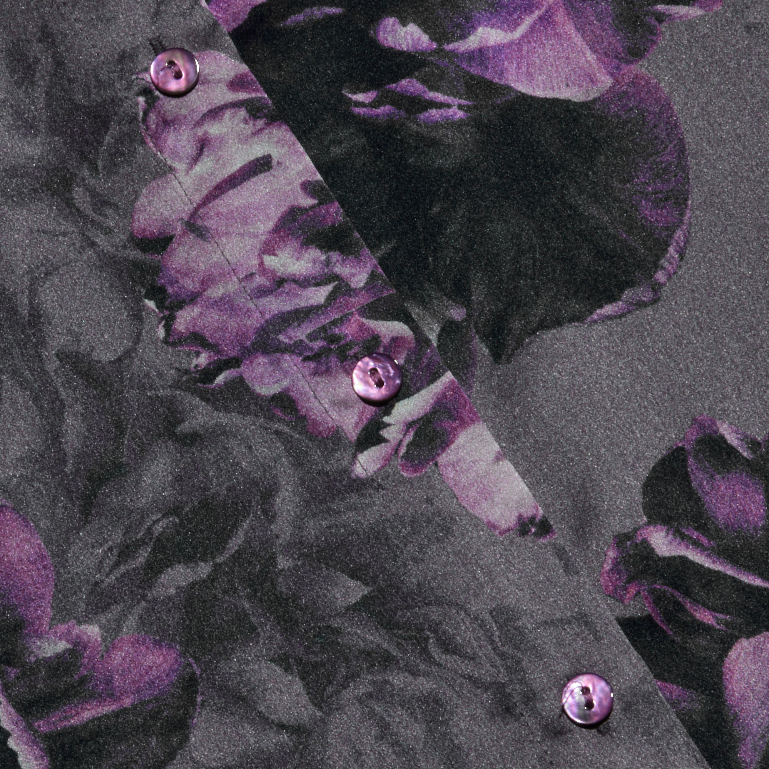 Close-up of the Tyler Silk Blouse by L’AGENCE, showcasing its purple floral print on a dark background and a diagonal row of glossy purple buttons.