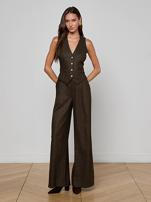 A woman stands indoors against a light gray wall, wearing the L'AGENCE Drina Pinstriped Vest with gold buttons and matching wide-legged trousers. She has long wavy brown hair and brown pointed-toe shoes.