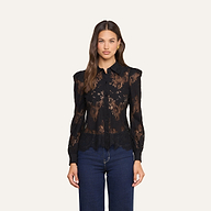 Una donna dai lunghi capelli castani indossa la camicetta Jenica Lace, un modello femminile in pizzo nero con bottoni e maniche lunghe, abbinata a un paio di jeans blu scuro. È in piedi davanti a uno sfondo beige a tinta unita e guarda direttamente verso la fotocamera.