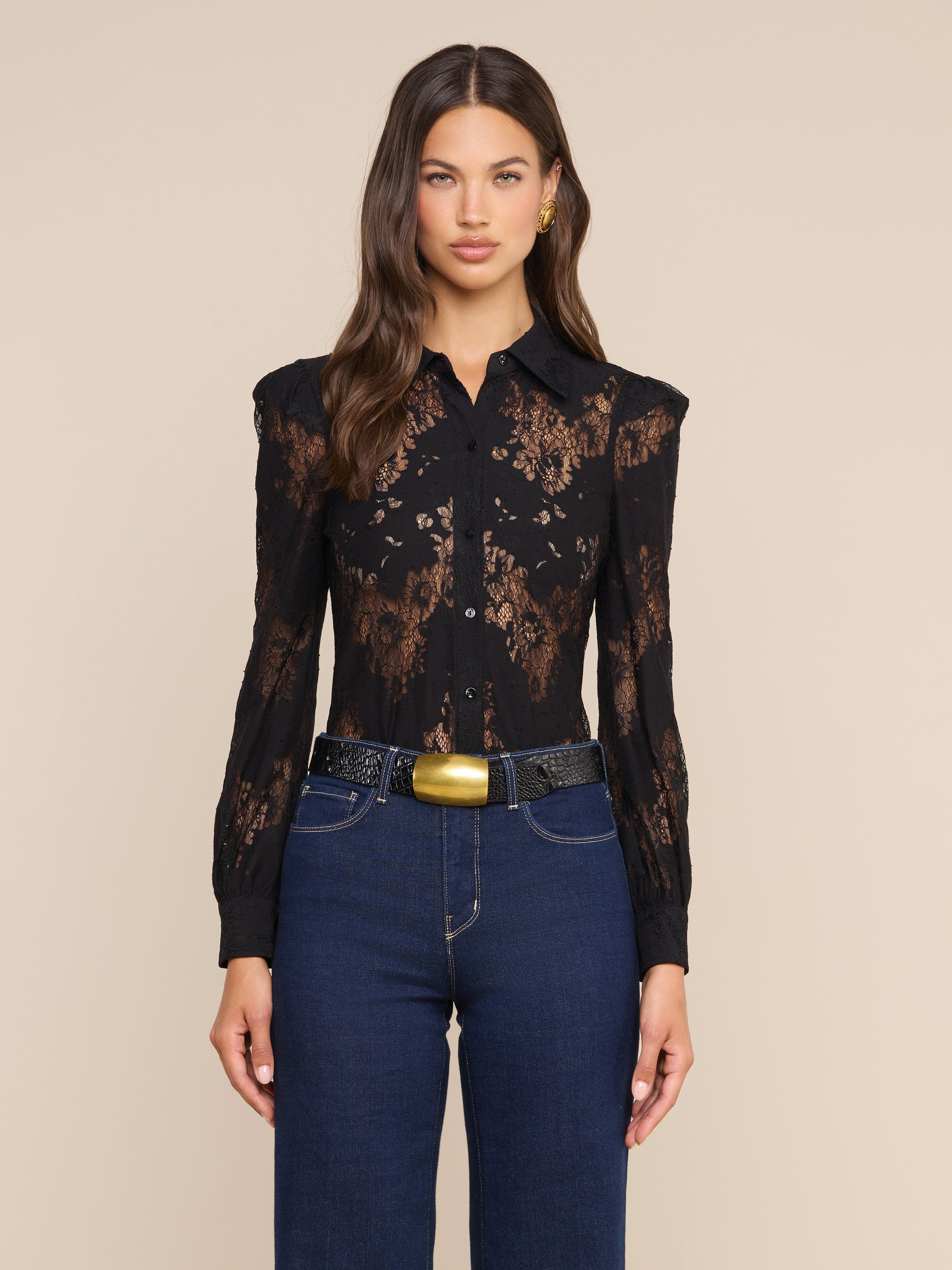 Una donna dai lunghi capelli castani indossa la camicetta in pizzo Jenica, abbinata a jeans blu a vita alta, una cintura nera con fibbia dorata e orecchini dorati, in posa su uno sfondo beige neutro.