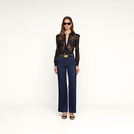 Una donna è in posa su uno sfondo beige e indossa la camicetta in pizzo Jenica, jeans blu scuro a vita alta, sandali neri con tacco, occhiali da sole e una cintura con fibbia dorata.
