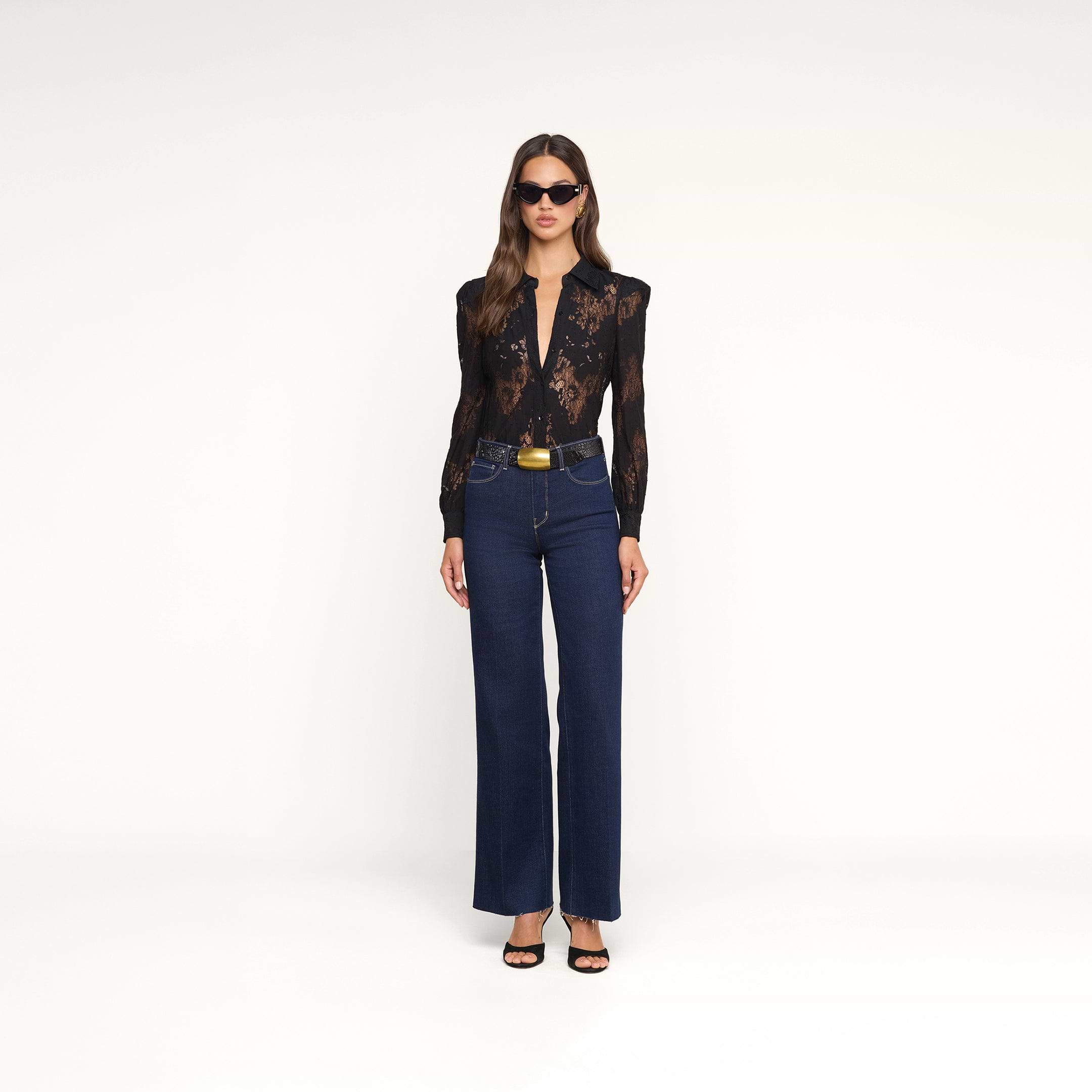 Una donna è in posa su uno sfondo beige e indossa la camicetta in pizzo Jenica, jeans blu scuro a vita alta, sandali neri con tacco, occhiali da sole e una cintura con fibbia dorata.