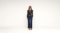 Woman wearing sunglasses, a sheer black long-sleeve top, dark blue high-waisted pants with a belt, and black heels stands against a plain white background.