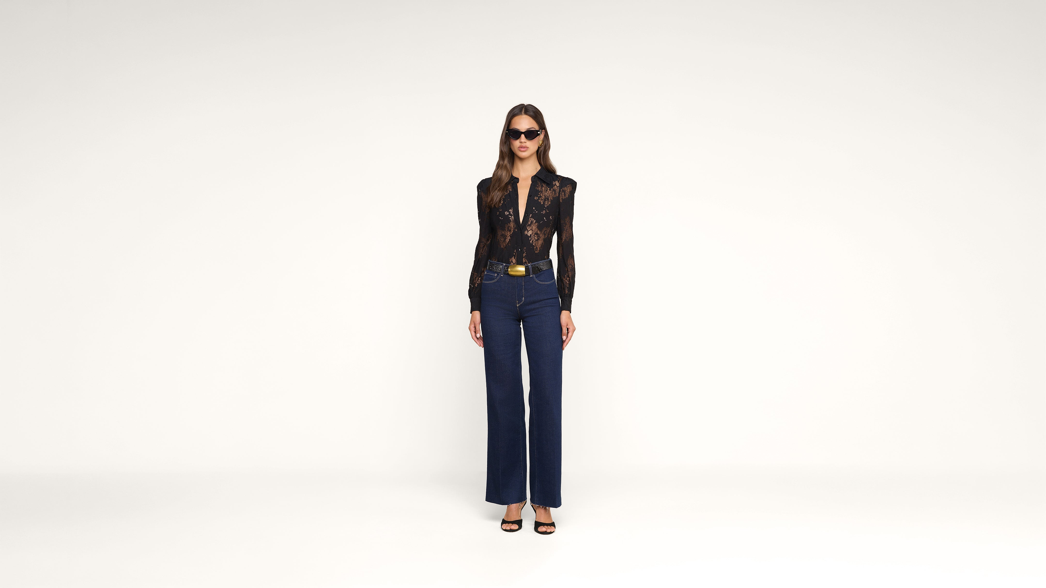 Woman wearing sunglasses, a sheer black long-sleeve top, dark blue high-waisted pants with a belt, and black heels stands against a plain white background.