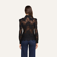 Una donna dai lunghi capelli castani, che indossa la camicetta Jenica Lace con spalle a sbuffo e jeans blu scuro, è in piedi di spalle alla fotocamera su uno sfondo beige.