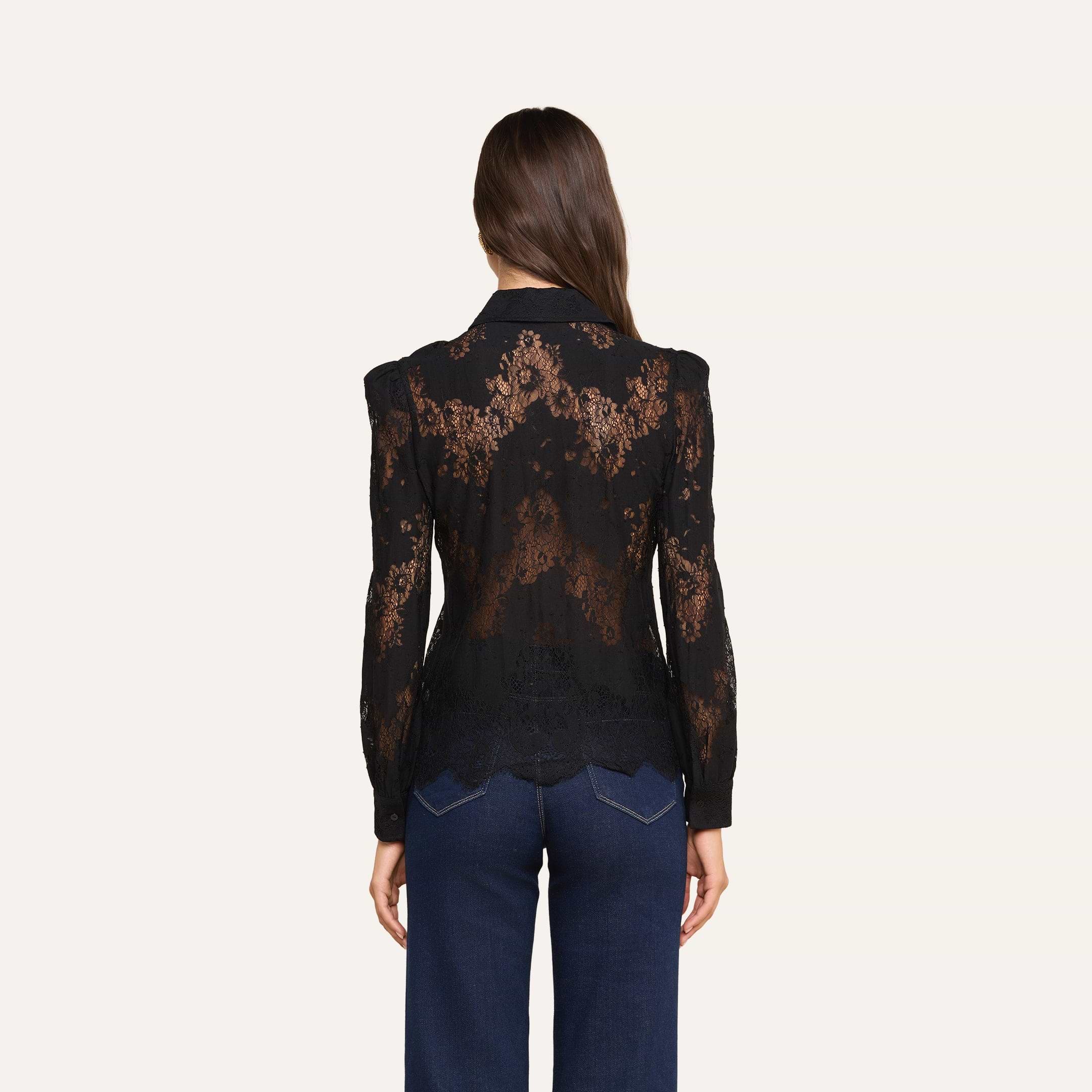 Una donna dai lunghi capelli castani, che indossa la camicetta Jenica Lace con spalle a sbuffo e jeans blu scuro, è in piedi di spalle alla fotocamera su uno sfondo beige.