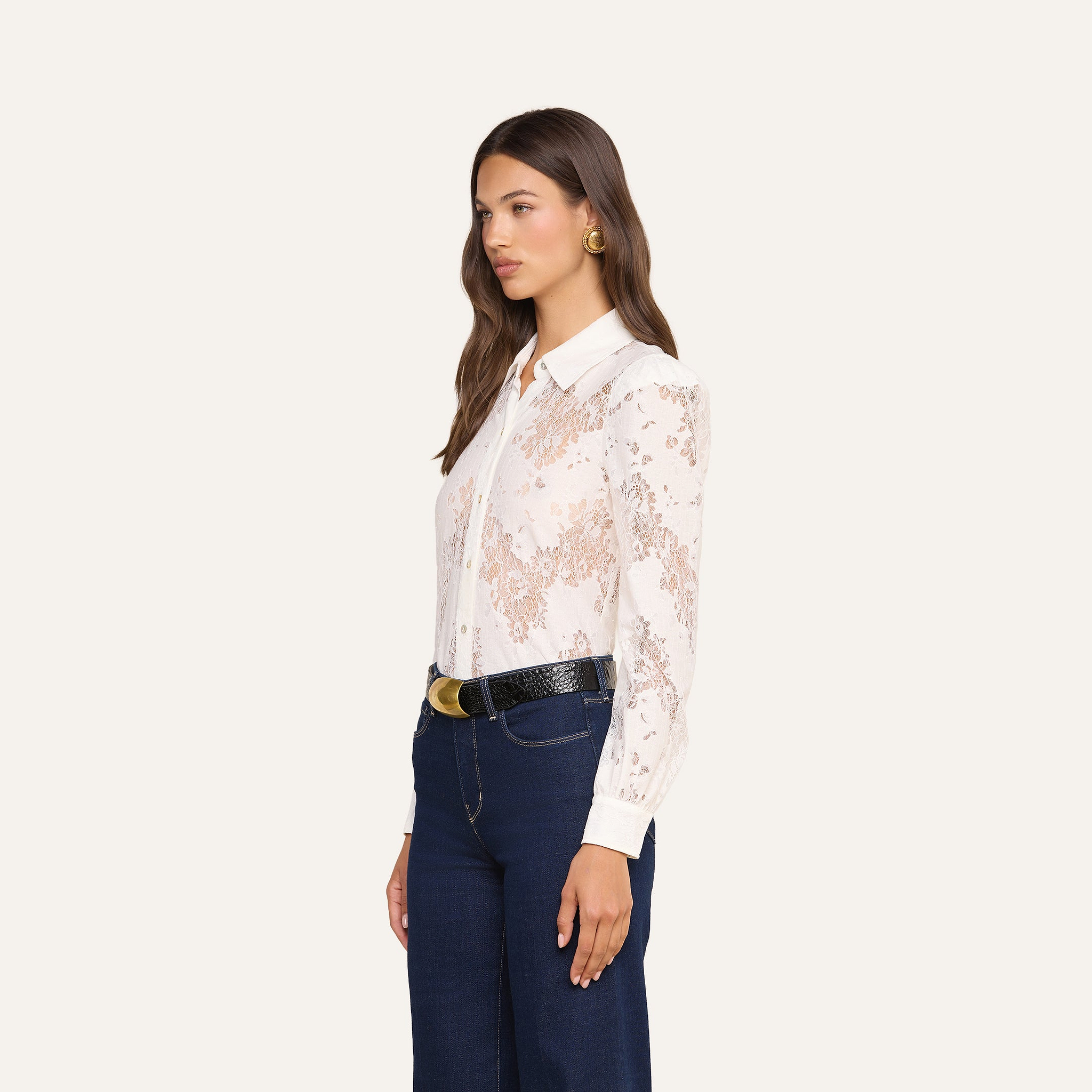 Woman standing sideways wearing the Jenica Lace Blouse—a white, long-sleeve cotton-blend floral lace top with puff shoulders—paired with dark blue pants, a black belt featuring a gold buckle, and gold hoop earrings against a plain light background.