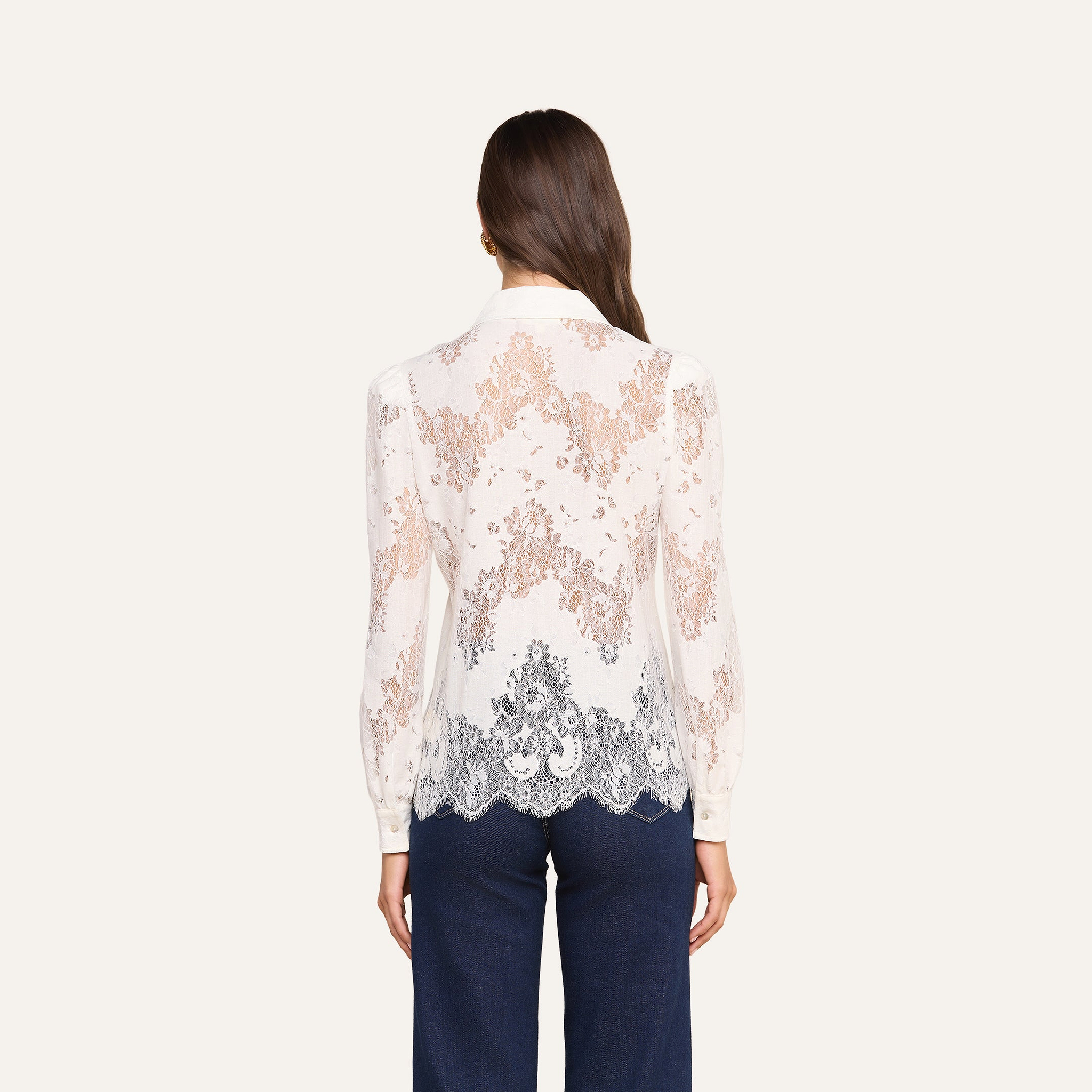 A woman stands with her back to the camera, wearing the Jenica Lace Blouse—a long-sleeve, sheer white floral lace top crafted from a cotton blend. The blouse features intricate patterns, scalloped edges, a collar, and subtle puff shoulders. She pairs it with dark blue pants against a plain, light-colored background.