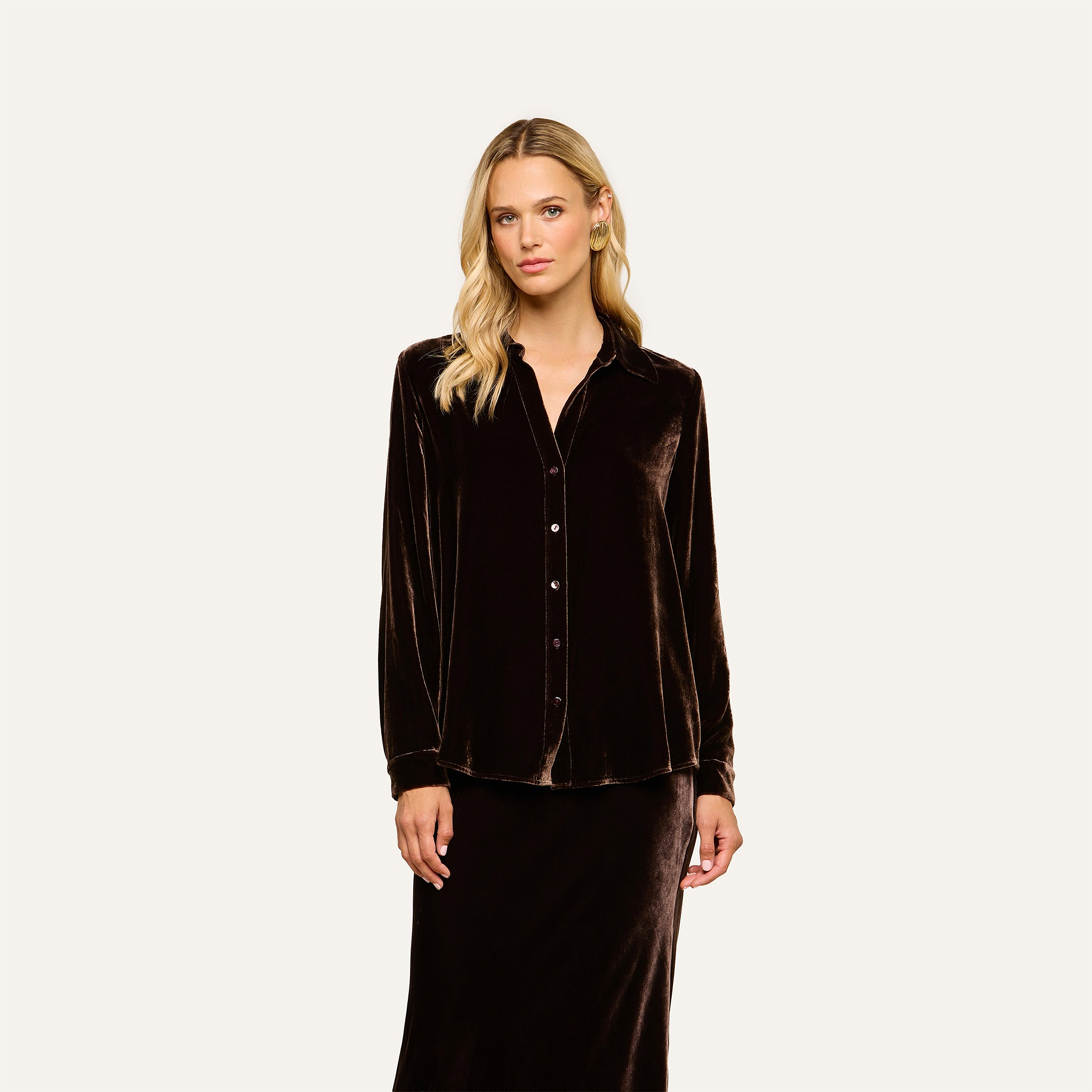 A woman with long blonde hair wears the Argo Velvet Blouse—a long sleeve button-down top in dark java brown velvet—paired with a matching skirt, standing against an off-white background.