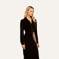A woman with long blonde hair wears the Argo Velvet Blouse in dark Java brown, featuring long sleeves. She stands against a plain light background, looking slightly to the side with a neutral expression.