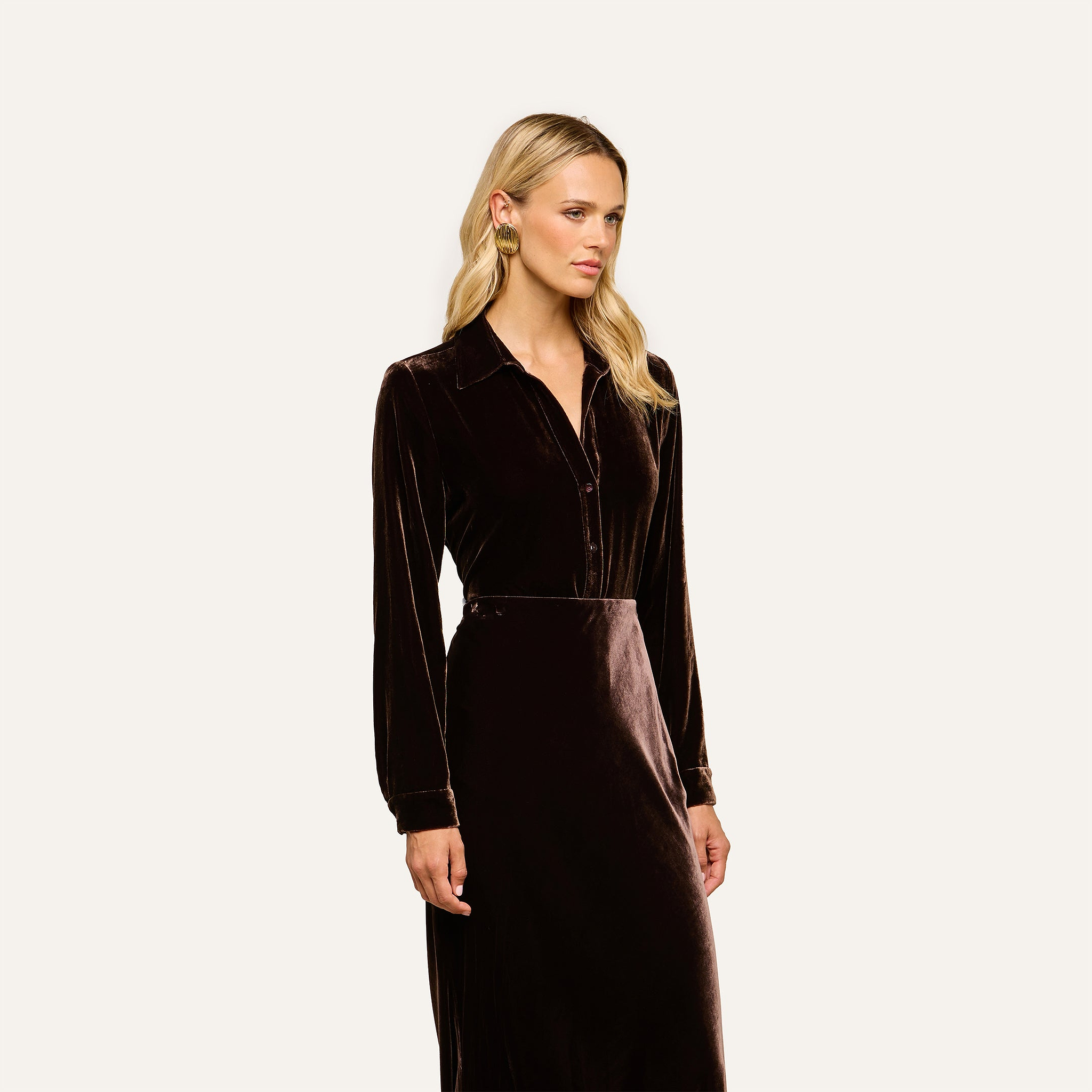 A woman with long blonde hair wears the Argo Velvet Blouse in dark Java brown, featuring long sleeves. She stands against a plain light background, looking slightly to the side with a neutral expression.