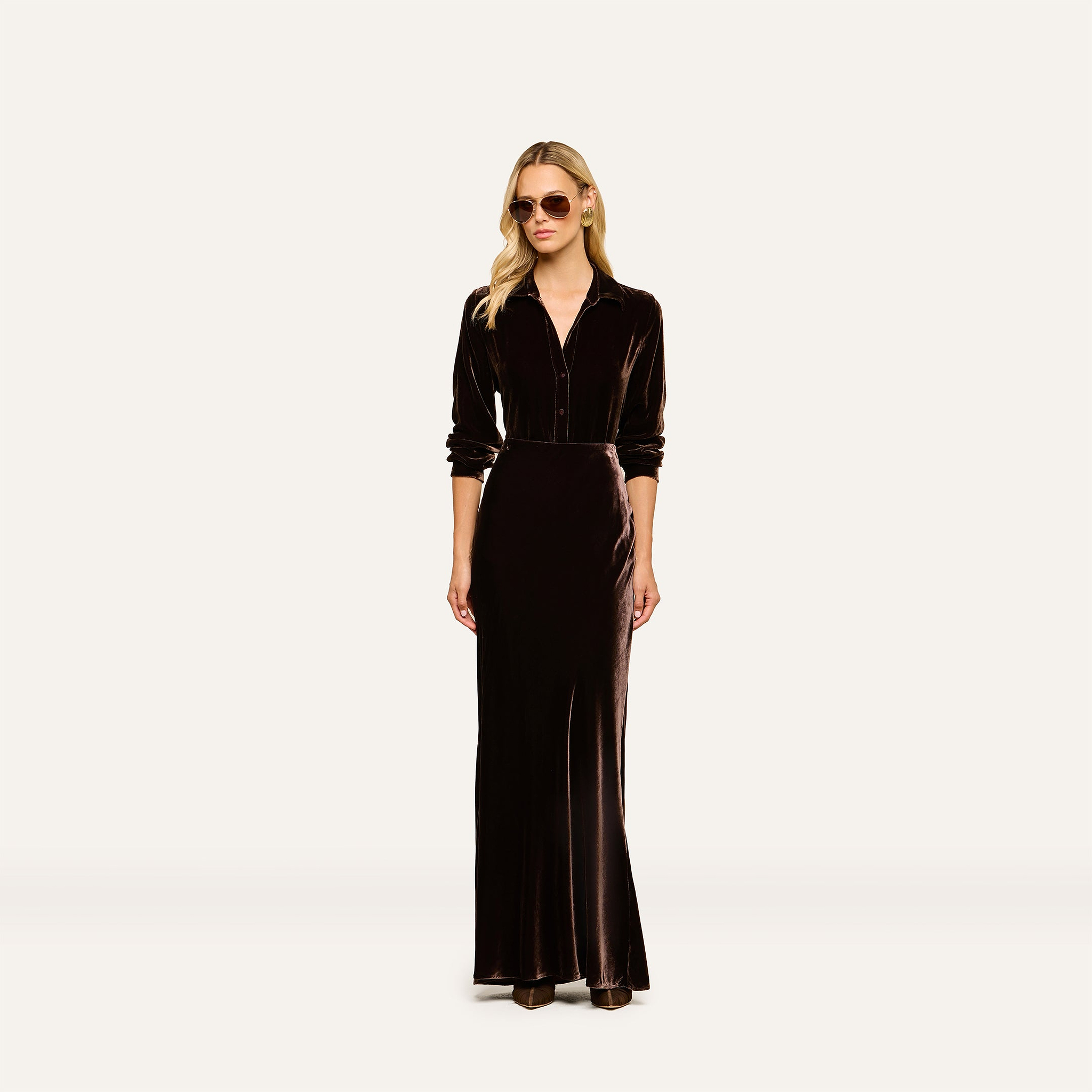 A woman wearing sunglasses and the Argo Velvet Blouse—a long sleeve, button-down top in dark java brown velvet—stands against a plain light background.